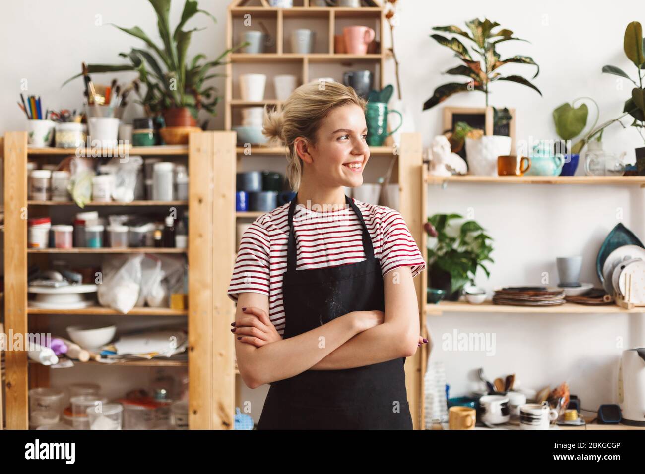 Chica sonriente en delantal negro y camiseta felizmente mirando a un lado con platos hechos a mano en estantes en el fondo en el estudio de cerámica Foto de stock