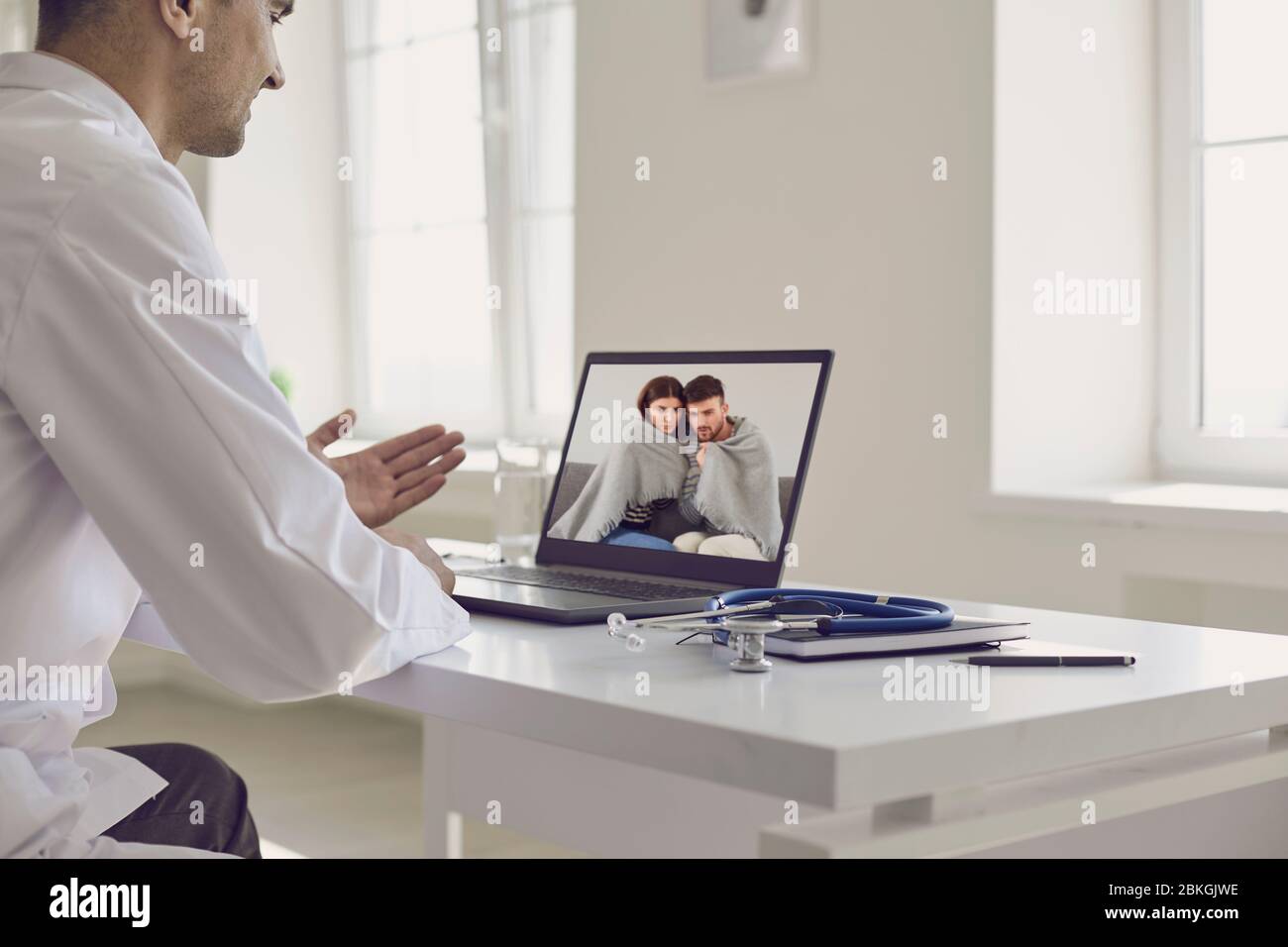Médico en línea. El médico habla con una pareja de pacientes usando un portátil sentado detrás de una pajita en una clínica. Consulta médica en línea. Foto de stock