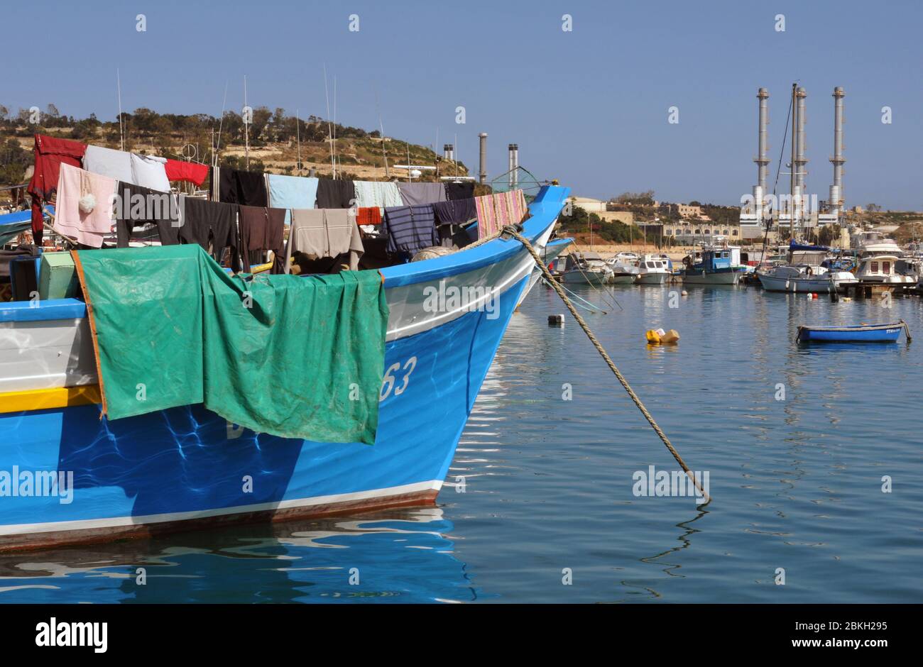 La lavandería se cuelga para secarse a bordo de un barco amarrado en el puerto en el pueblo de Marsaxlokk, Malta. La central eléctrica de Delimara está en segundo plano. Foto de stock