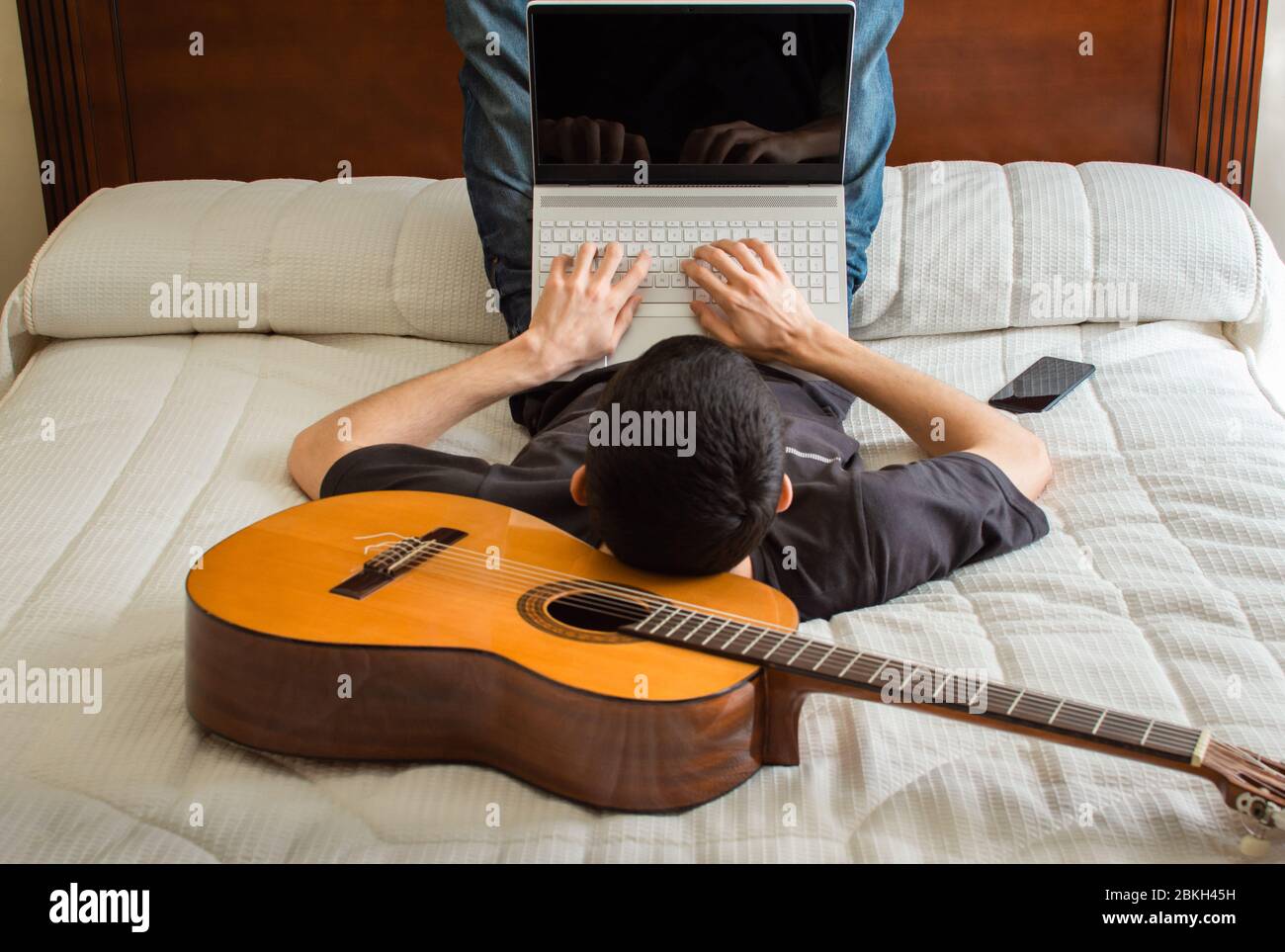 Hombre descansando en una cama y usando un ordenador portátil después de  tocar la guitarra en casa. Tiempo libre en casa: Tecnología y música.  Músico utilizando la guitarra como almohada Fotografía de