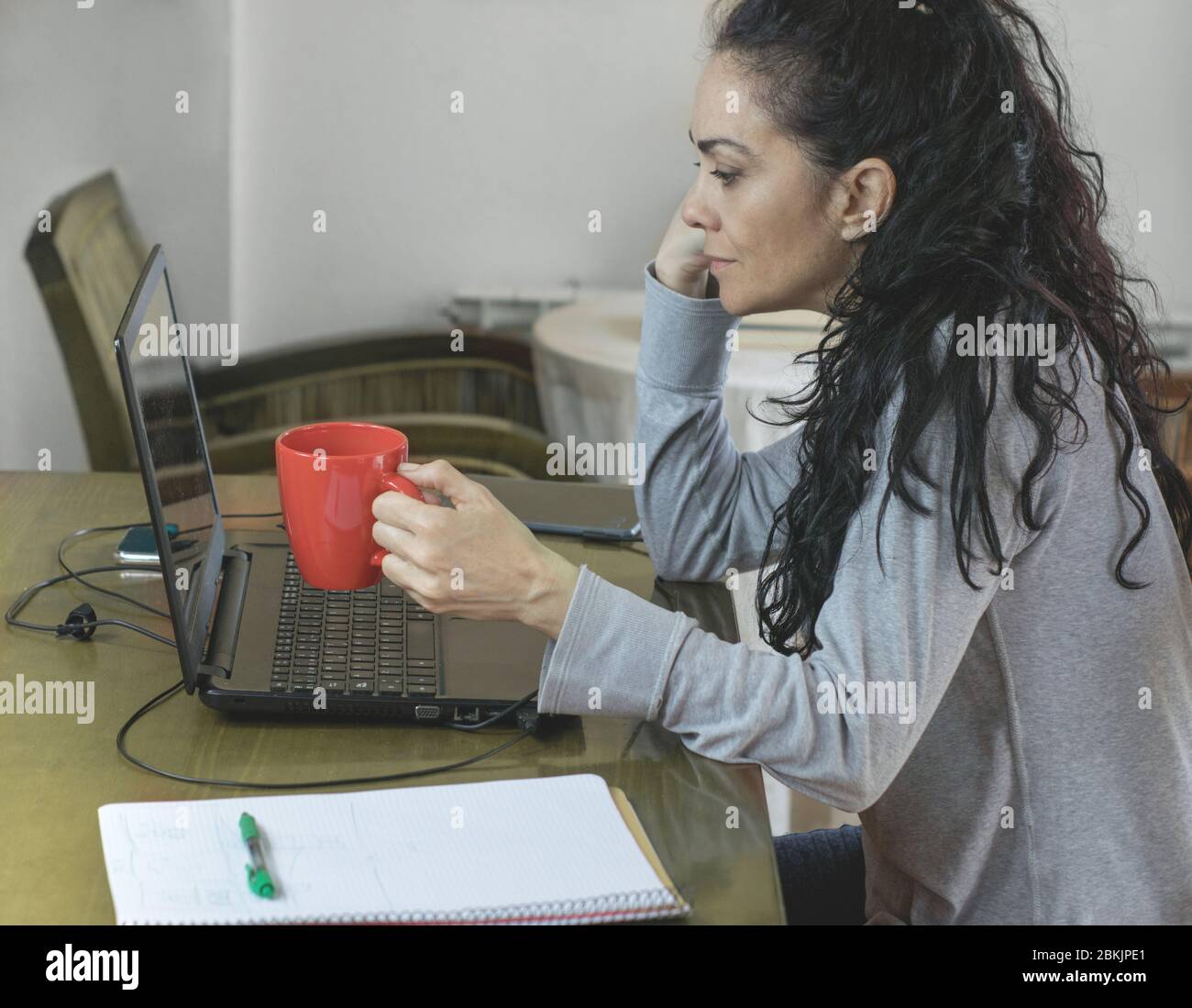 Mujer morena teletrabajo en casa para COVID-19, mirando el ordenador con una taza de café en la mano Foto de stock