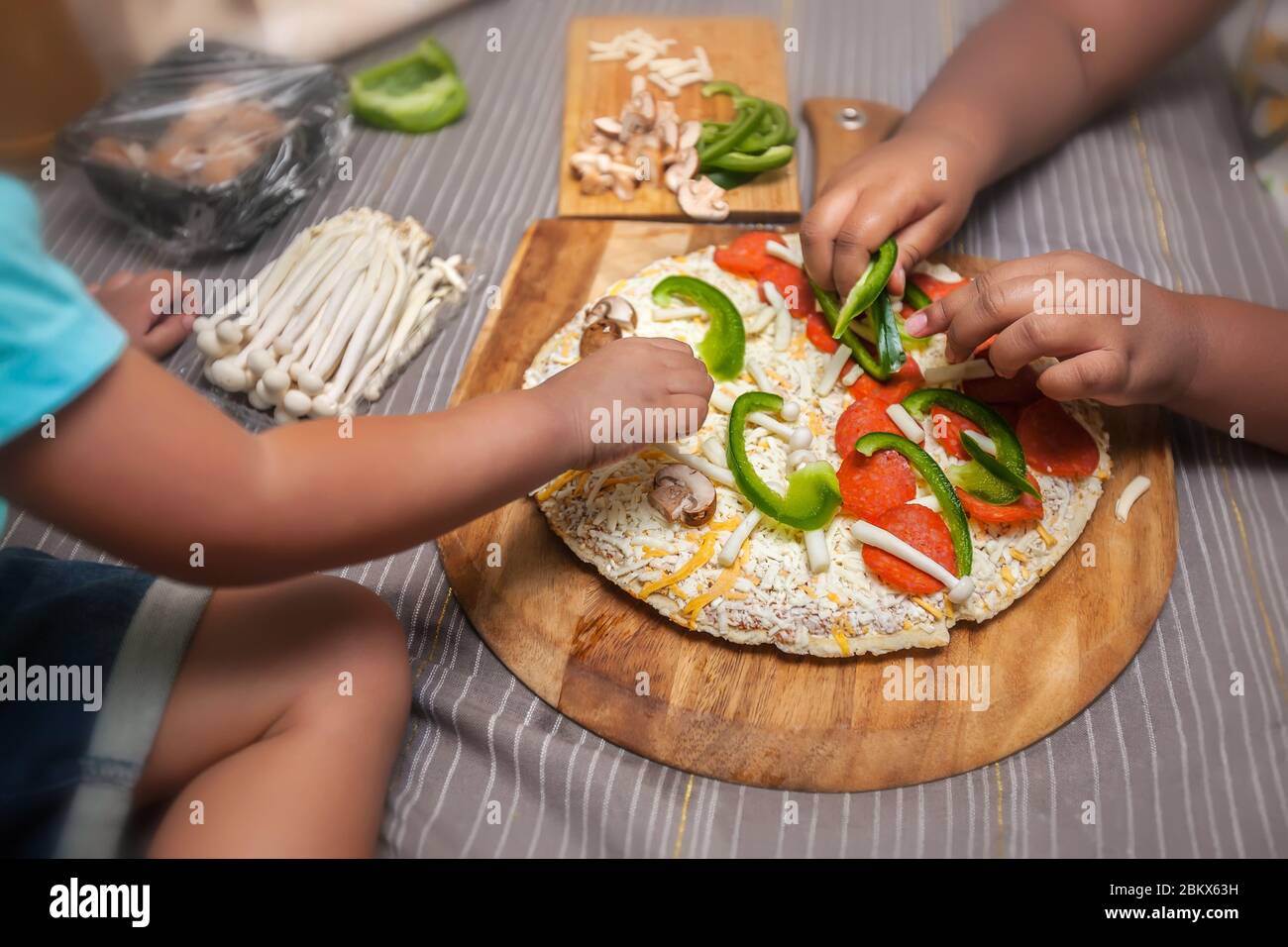 Los niños agregan ingredientes frescos a una pizza de queso congelado en una cáscara de pizza de madera. Foto de stock