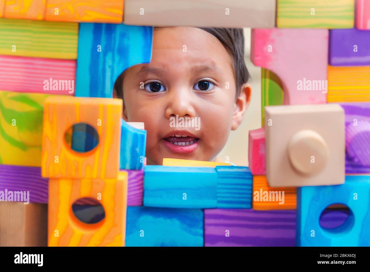 Un niño pequeño mostrando su cara a través de una ventana desde detrás de una pared de bloques de construcción con varias formas y colores. Foto de stock