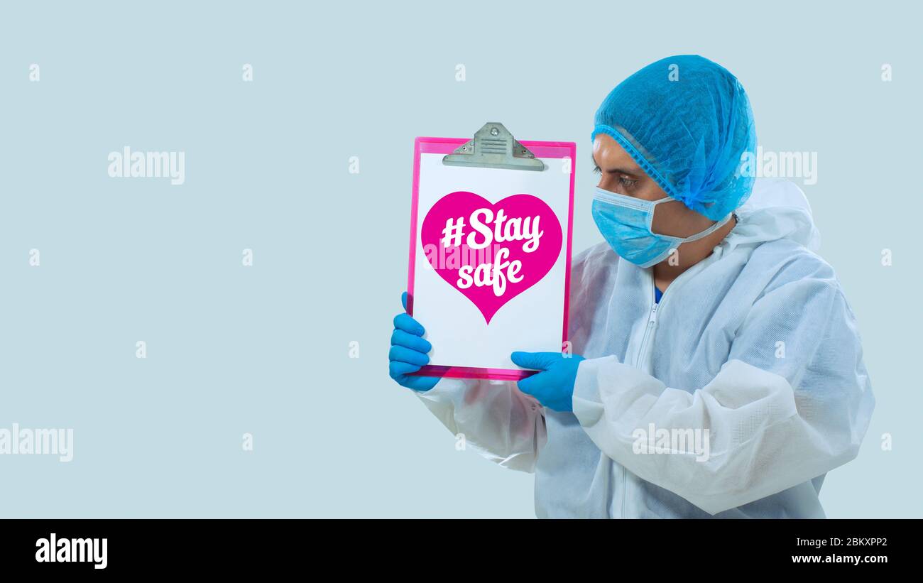 Médico con máscara y guantes azules sosteniendo una pizarra blanca con el mensaje: MANTÉNGASE SEGURO, dentro de un corazón rosa sobre fondo blanco Foto de stock