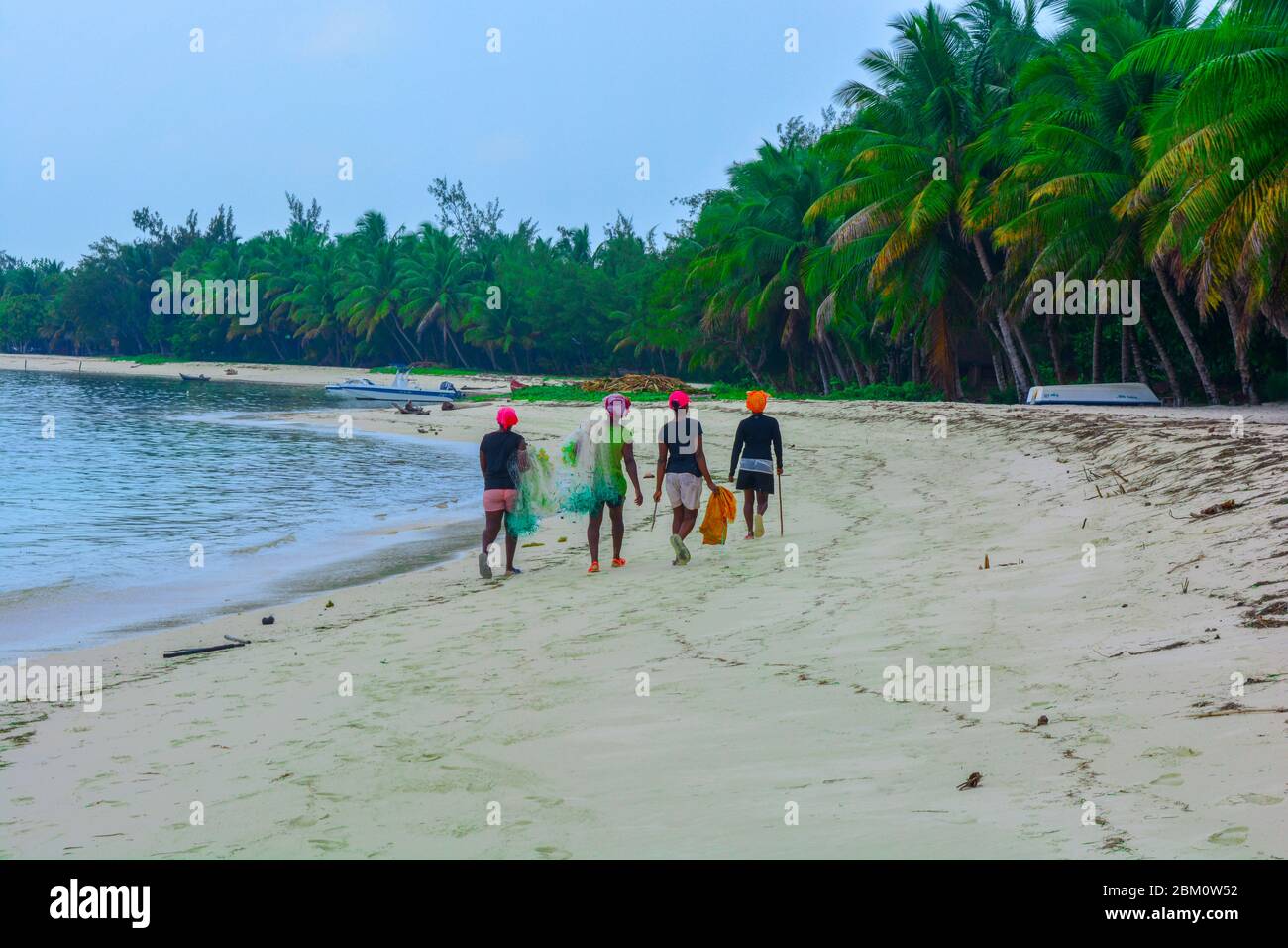 Cuatro mujeres africanas negras caminando en una fantástica playa con una larga línea de palmeras verdes. Pescadores malgaches con redes de pesca y sombreros coloridos Foto de stock