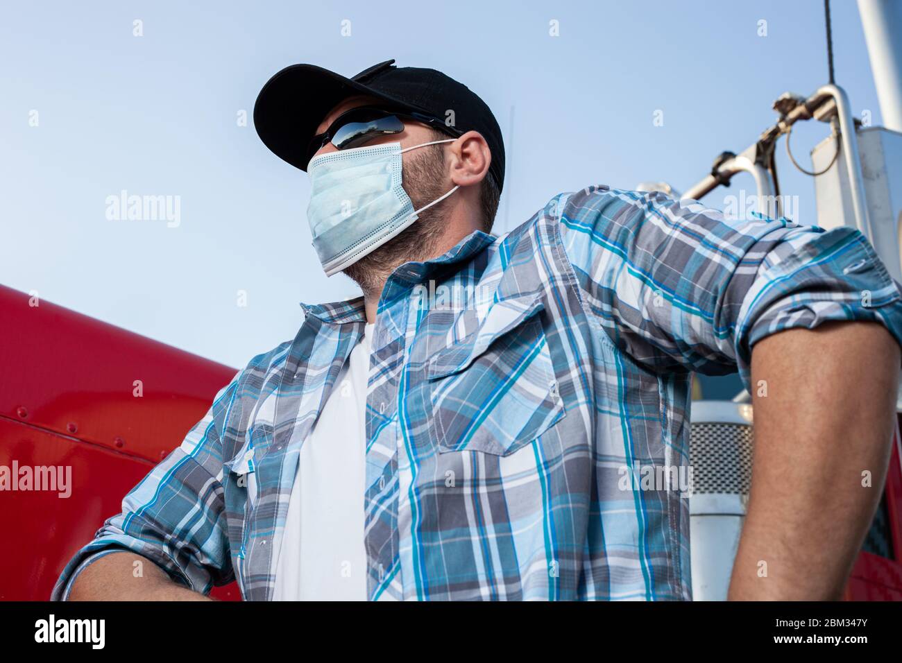 Conductor de camión de aspecto fresco vestido con ropa informal con camisa a cuadros y gorra negra de béisbol lleva máscara médica protectora. Foto de stock
