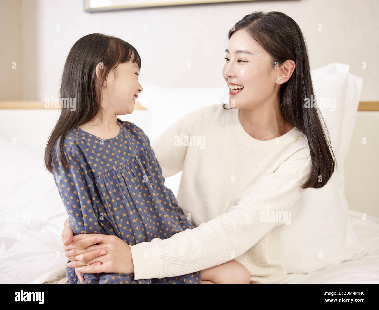 madre asiática joven y hija de edad elemental sentada en la cama relajándose jugando charlando en casa Foto de stock