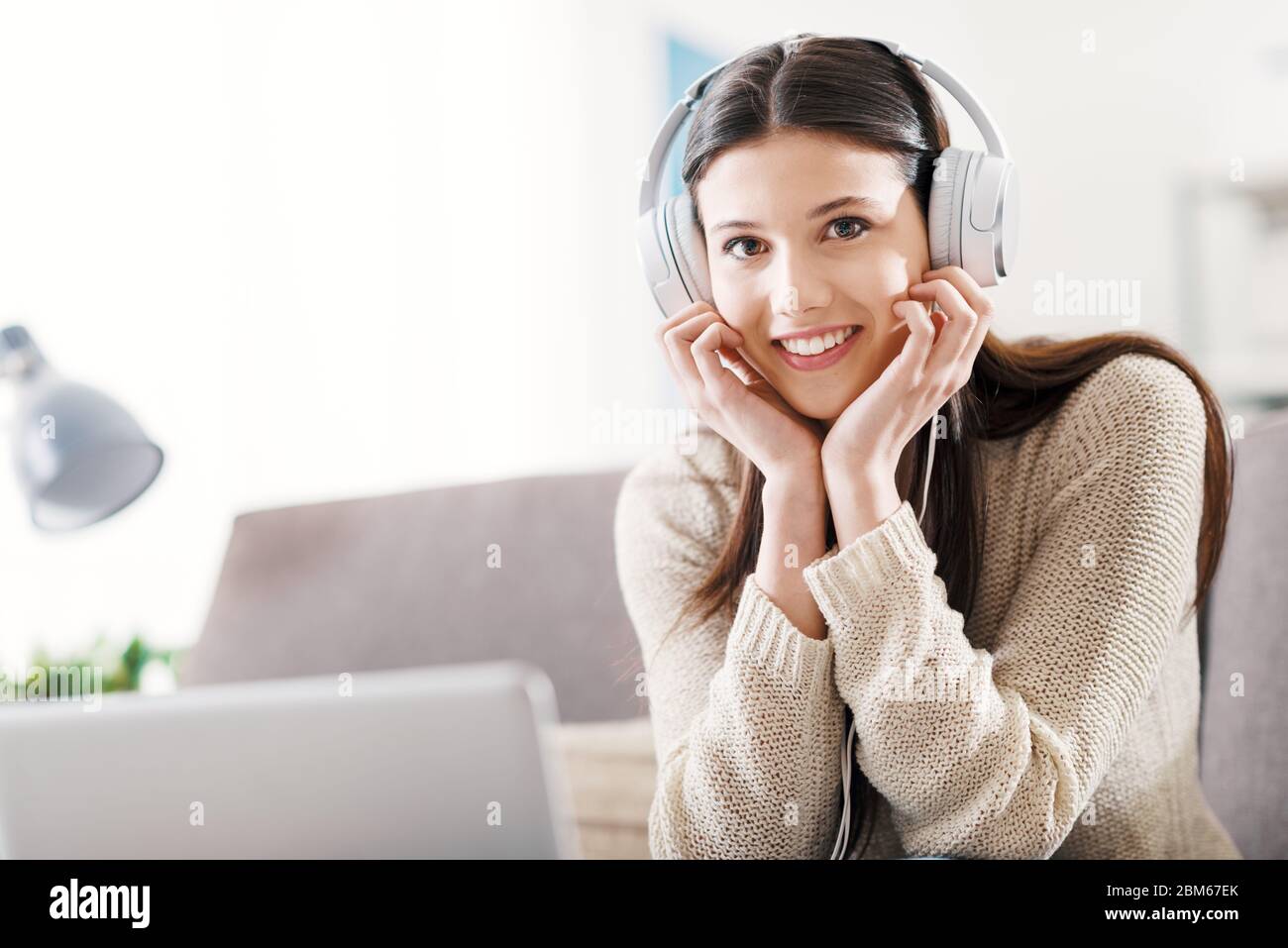 Joven mujer sonriente en casa relajándose en el sofá, está usando un ordenador portátil y escuchando música con auriculares Foto de stock