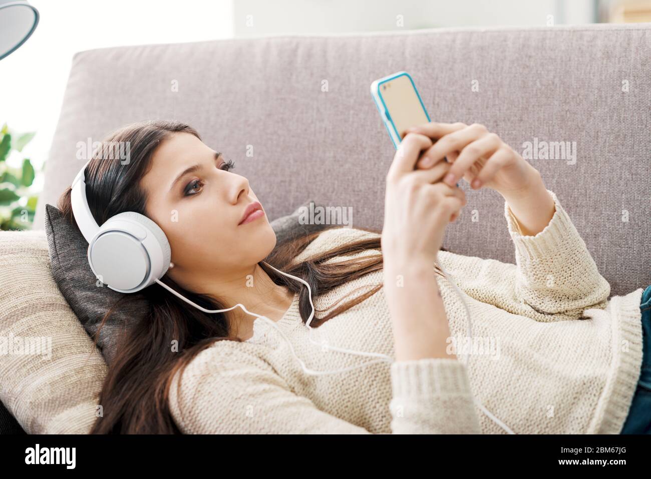 Atractiva chica relajado en el sofá y utilizar aplicaciones en su smartphone, ella está usando audífonos Foto de stock