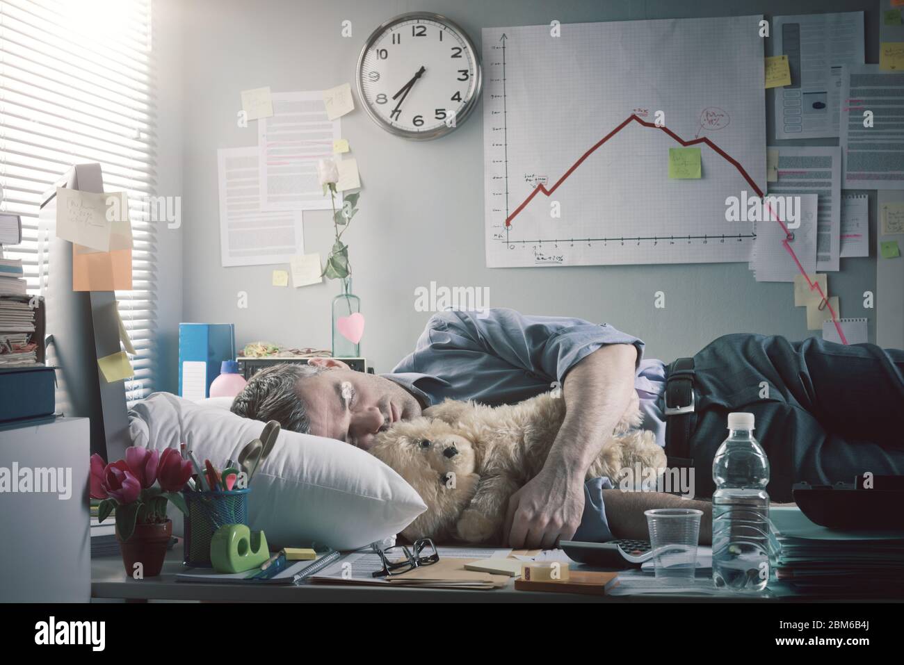 Trabajador de oficina divertido durmiendo en la oficina durante la noche con oso de peluche. Foto de stock