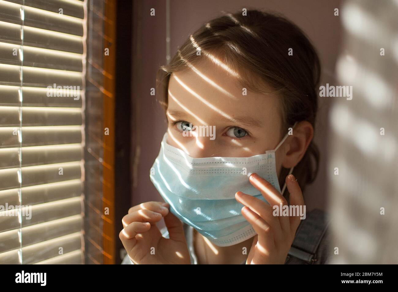 La niña está sentada en el alféizar de la ventana y tratando de poner una máscara desechable protectora en su cara durante el auto-aislamiento en casa debido al Coronavirus Foto de stock