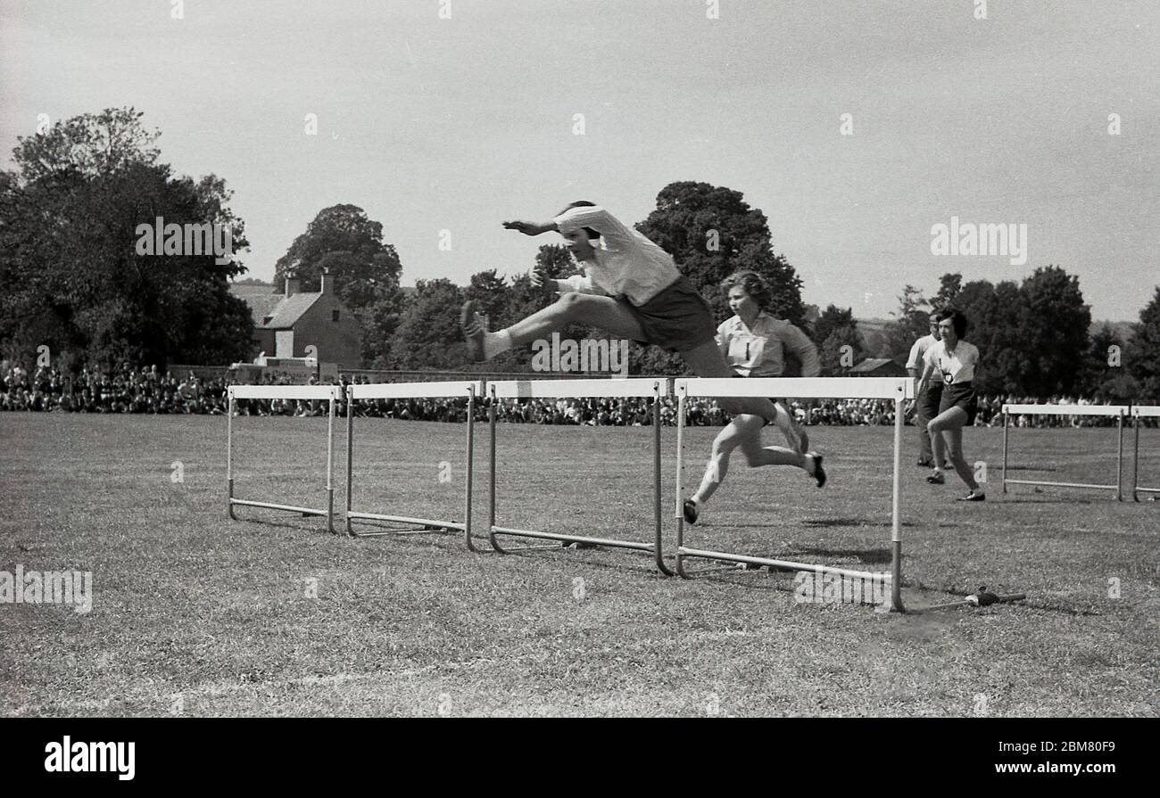 Deportes de secundaria, Inglaterra, c1960, fuera en una pista de hierba, las adolescentes compiten en una carrera de obstáculos, con una niña en el frente saltando sobre una barrera o obstáculo mostrando una buena técnica. Foto de stock