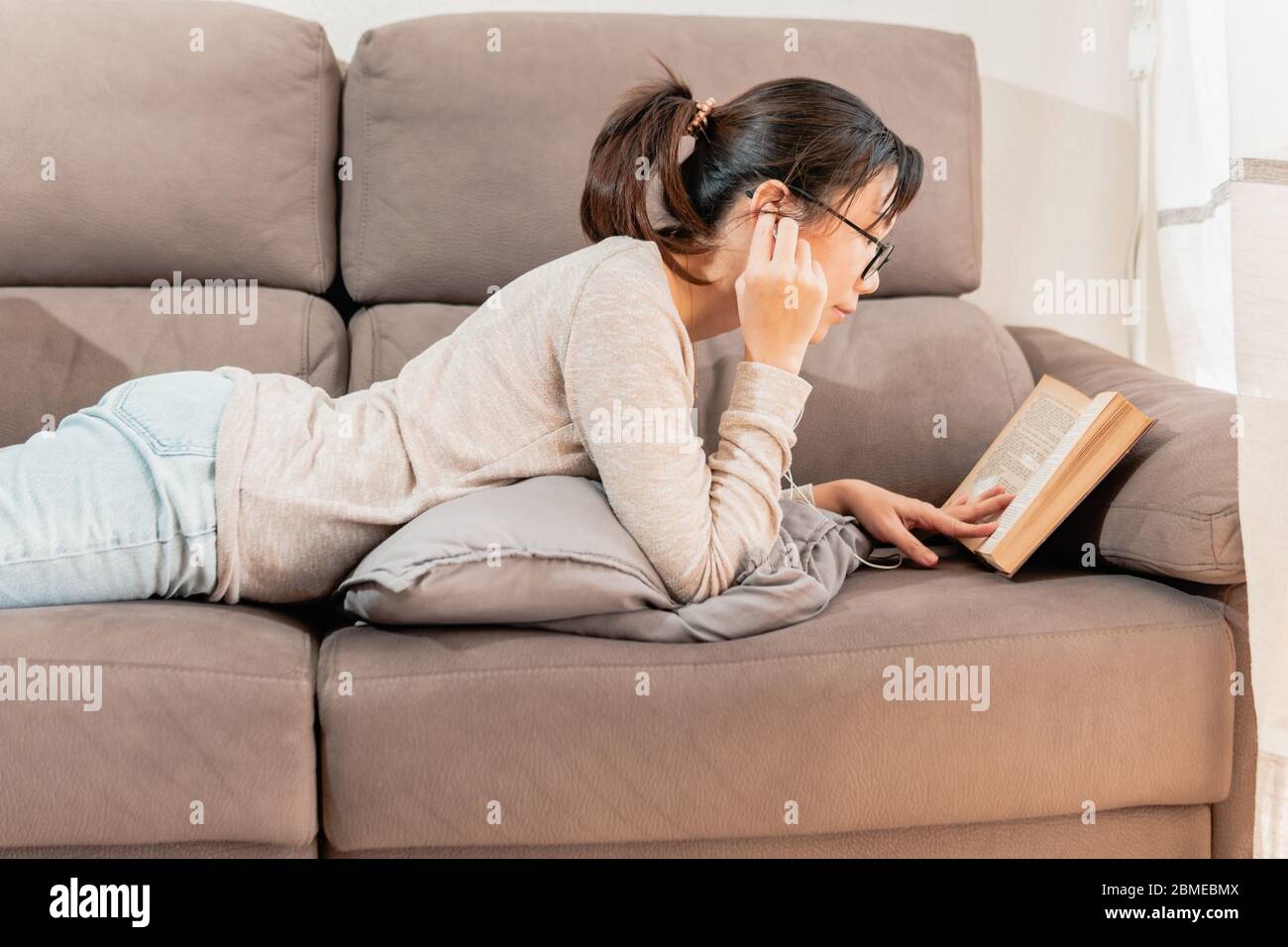 Mujer asiática con gafas leyendo un libro y escuchando música acostada en el sofá con los pies hacia arriba. Niña milenaria leyendo un viejo libro en casa. Foto de stock