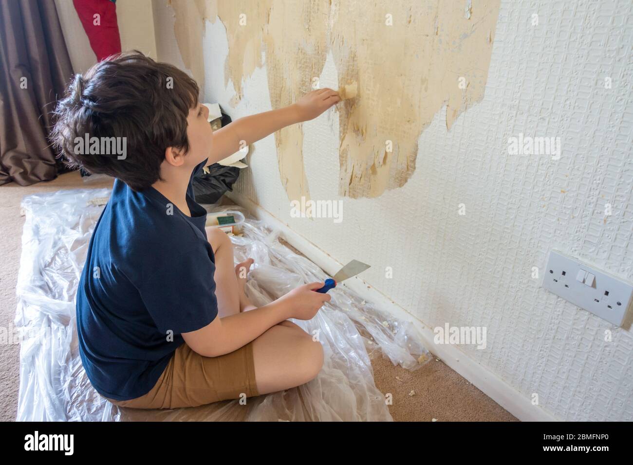 Un niño ayuda a quitar el papel pintado viejo de una pared como la primera etapa de redecorar una habitación. Foto de stock