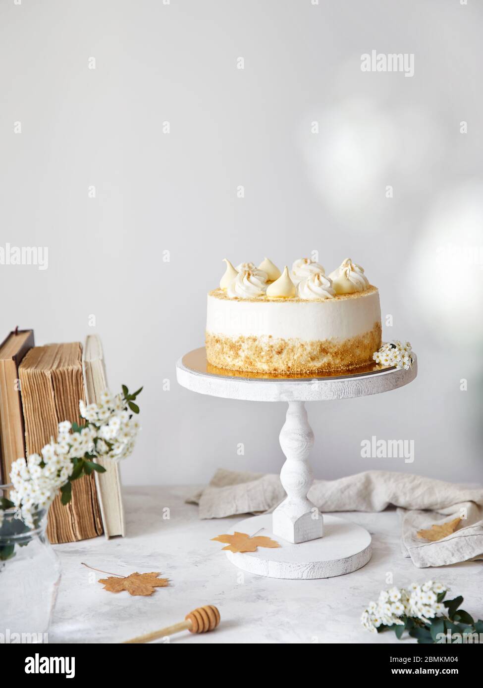 Pastel ruso casero con miel Medovik con crema y merengue en la parte superior. Flores blancas, hojas de otoño, libros viejos cerca en la mesa blanca Foto de stock