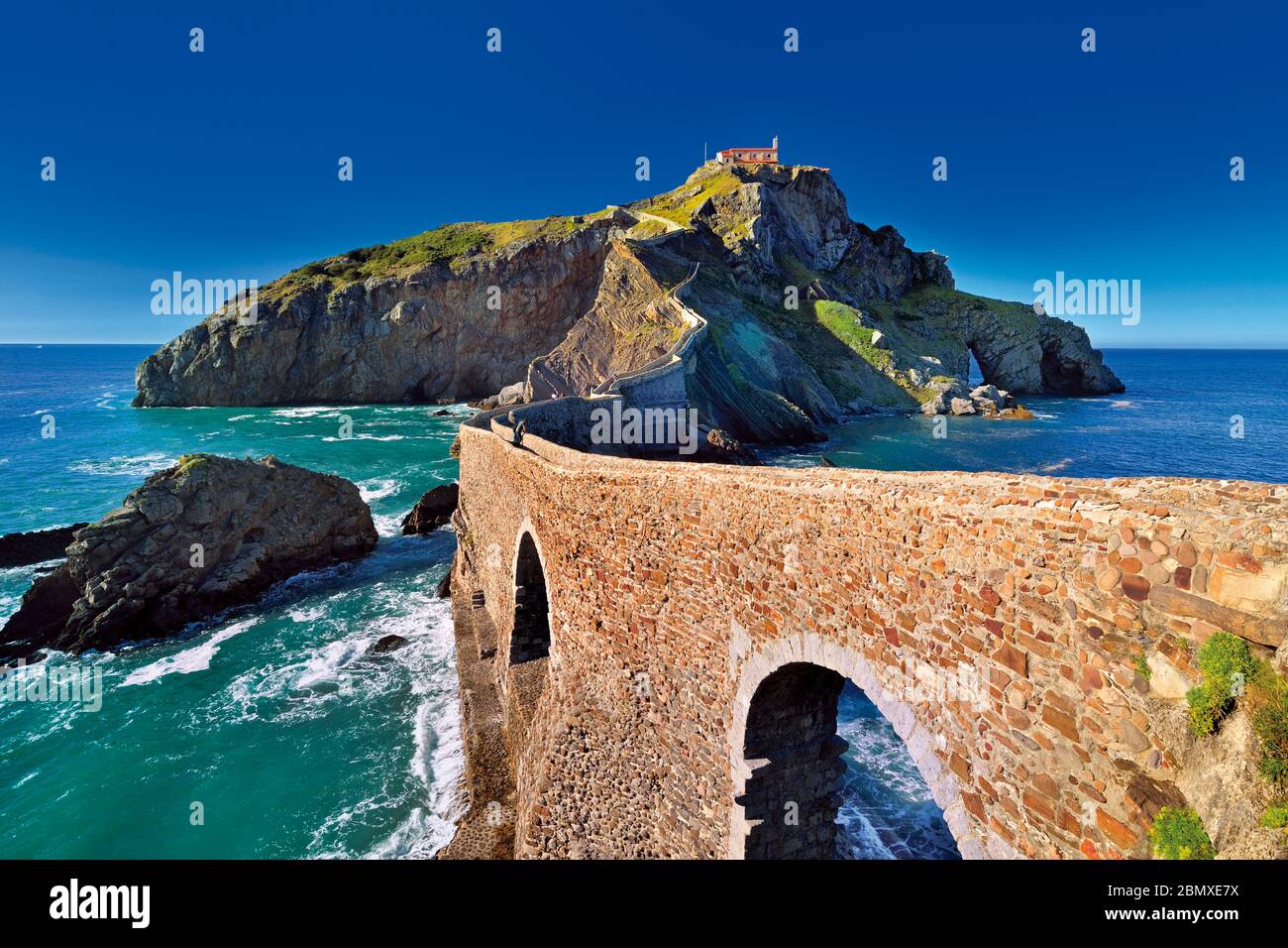 Puente de piedra arcada que conduce a una pequeña isla rocosa con una pequeña capilla en la parte superior Foto de stock