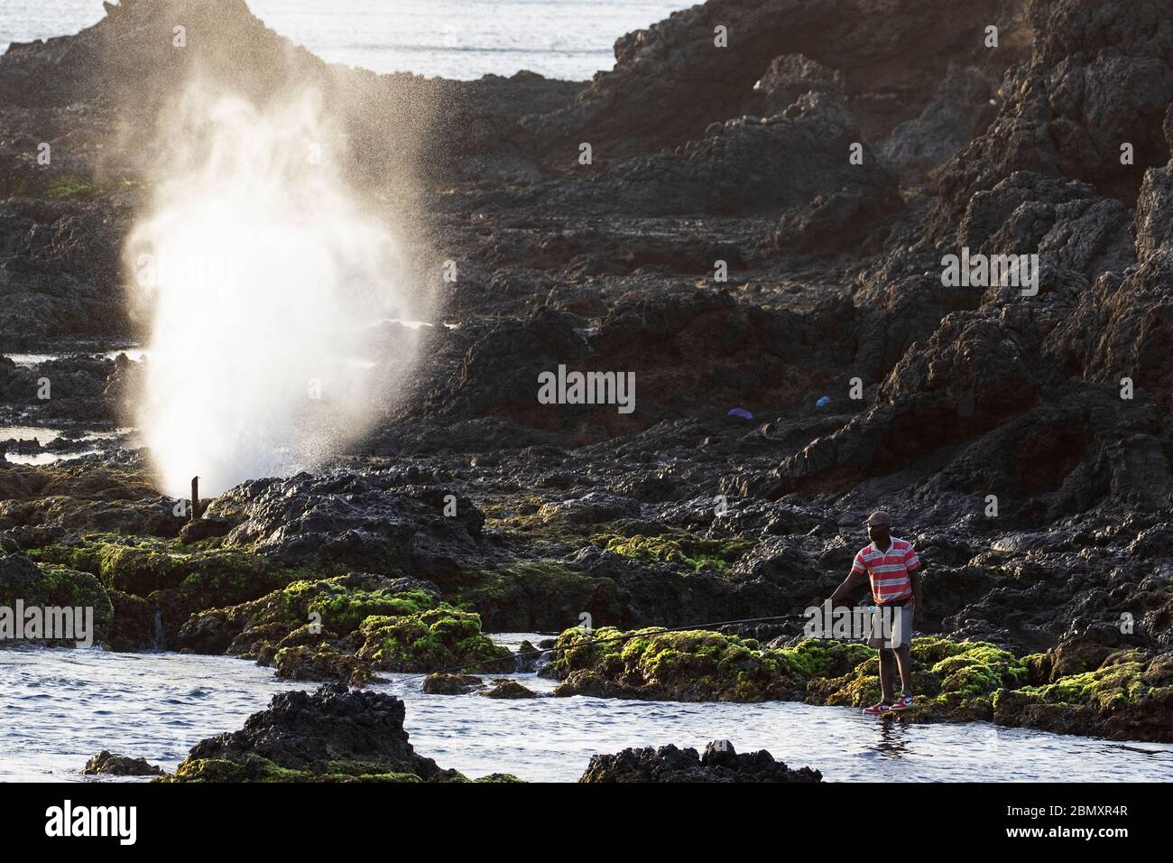 Pescador y olas rompiendo en rocas volcánicas en el surf en la punta Ponta Temerosa en la isla Santiago, Cabo Verde / Cabo Verde Foto de stock