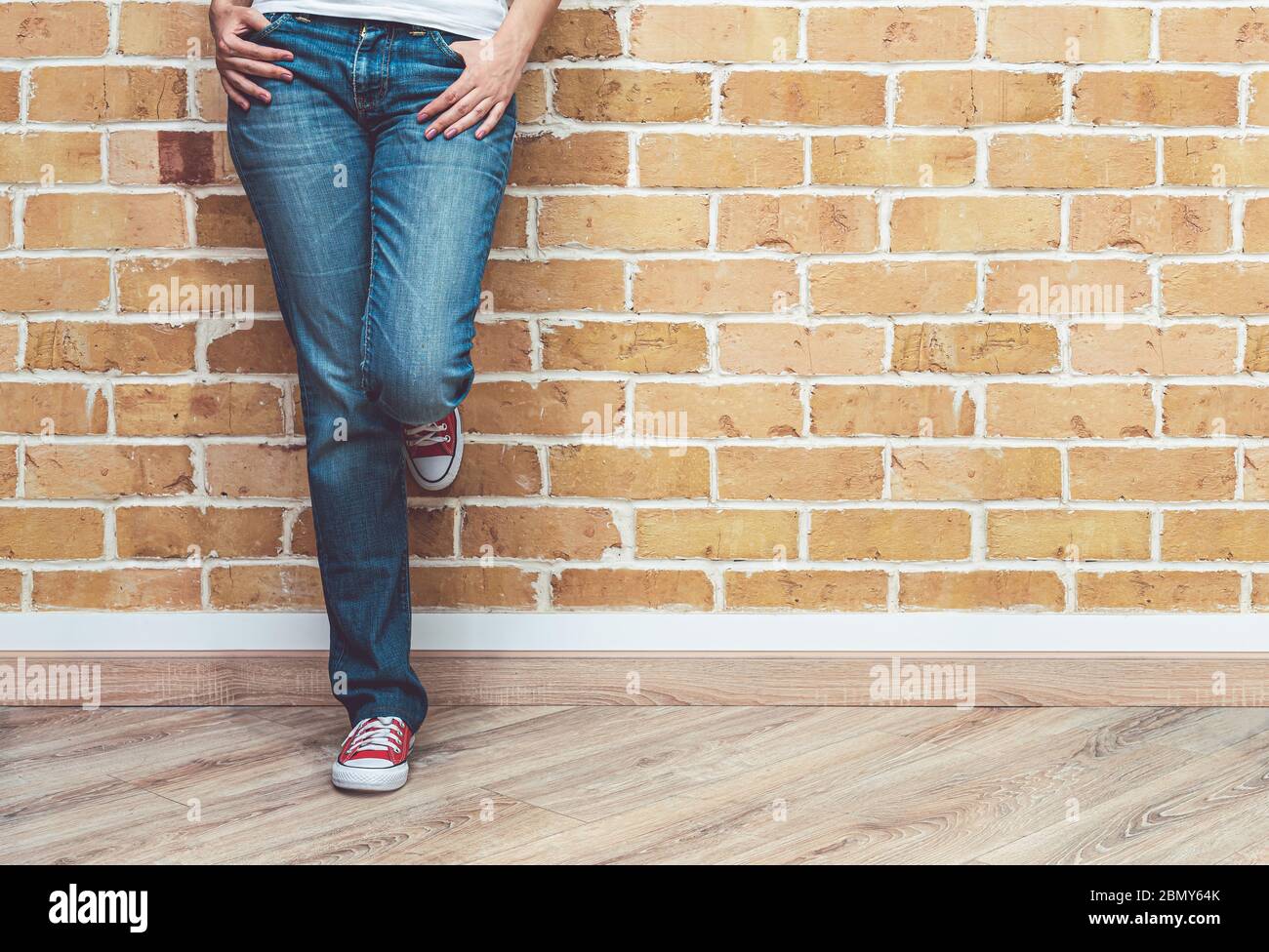 Mujer piernas en jeans y zapatillas de deporte rojas, mujer de pie frente la pared de ladrillo, concepto de Fotografía de -
