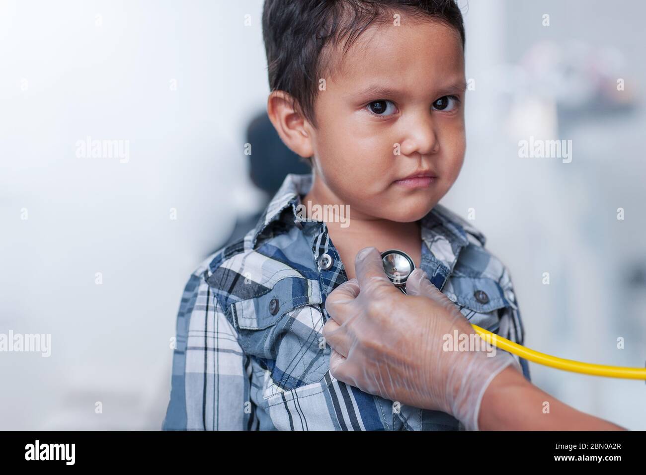 Un niño en edad preescolar que recibe una prueba de detección del corazón, usando un estetoscopio en el pecho con una camisa de abajo con botones. Foto de stock
