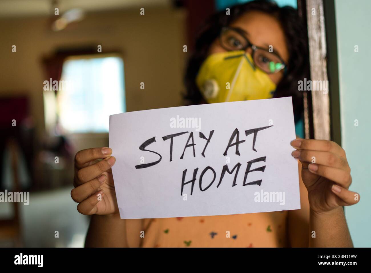 Una niña India que lleva una máscara de cara sostiene un cartel en las manos mostrando un mensaje "hay en casa" durante la pandemia de COVID-19 para mantener el distanciamiento social. Foto de stock