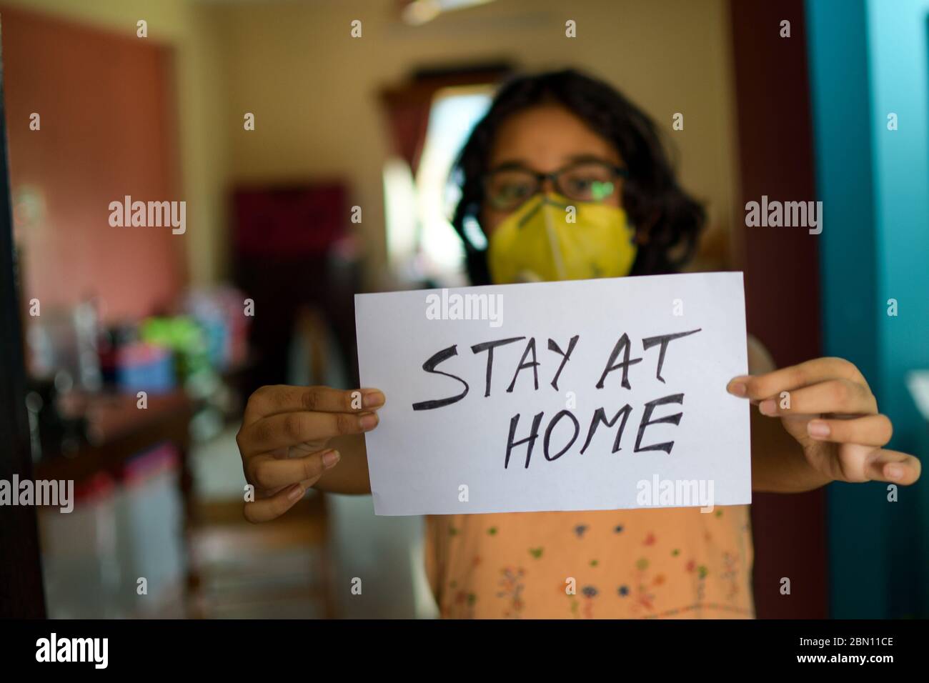 Una niña India que lleva una máscara de cara sostiene un cartel en las manos mostrando un mensaje "hay en casa" durante la pandemia de COVID-19 para mantener el distanciamiento social. Foto de stock