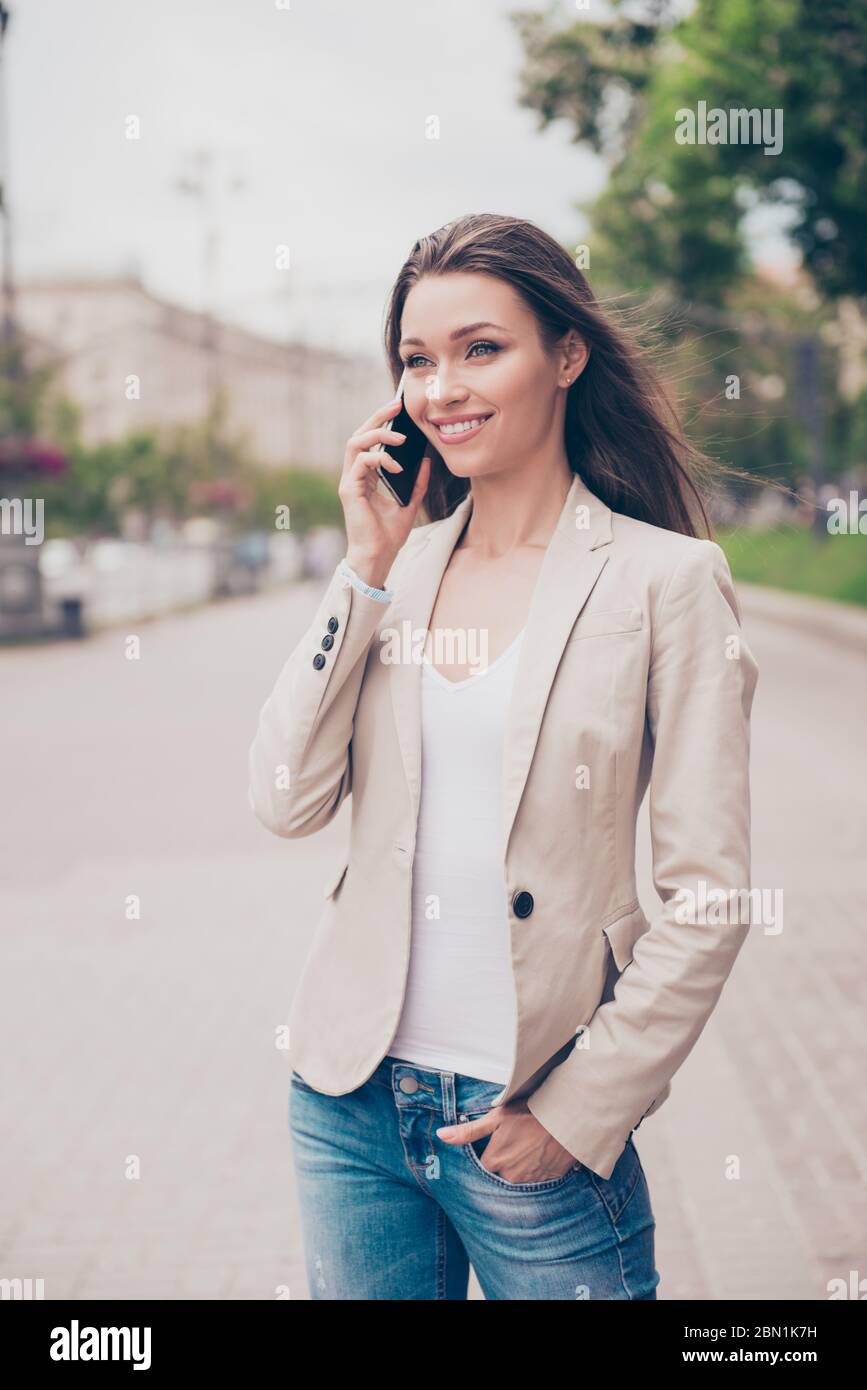 Joven bonita dama en un traje formal es talikg en el móvil, sonriendo. Ella  está en una caminata de primavera afuera, disfrutando, sosteniendo mano en  el bolsillo de jeans Fotografía de stock -