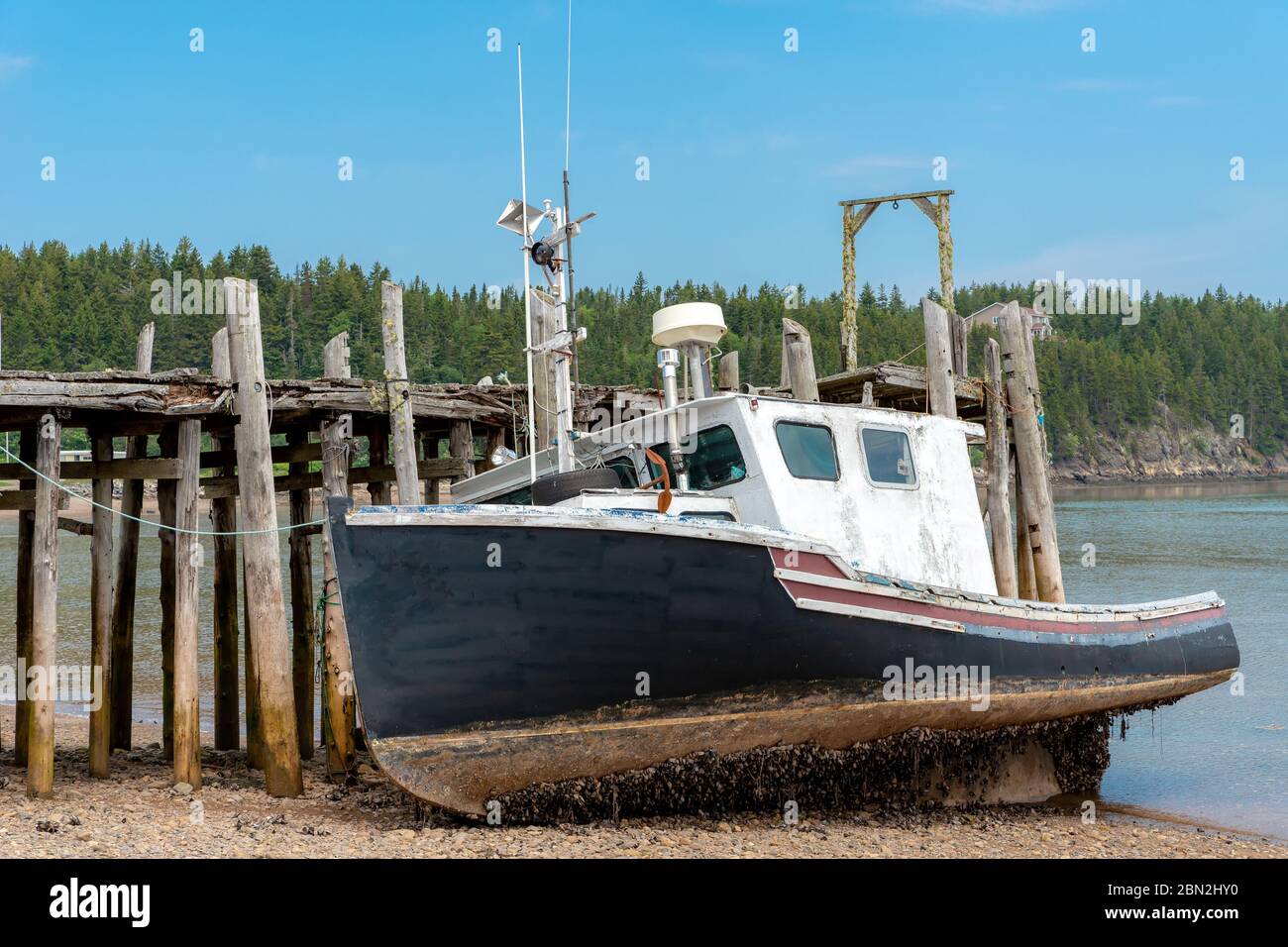 Un barco de pesca no identificado en marea baja. Se encuentra en una playa junto a un antiguo muelle de madera. Mano de los percebes desde abajo. Cielo azul arriba. Foto de stock
