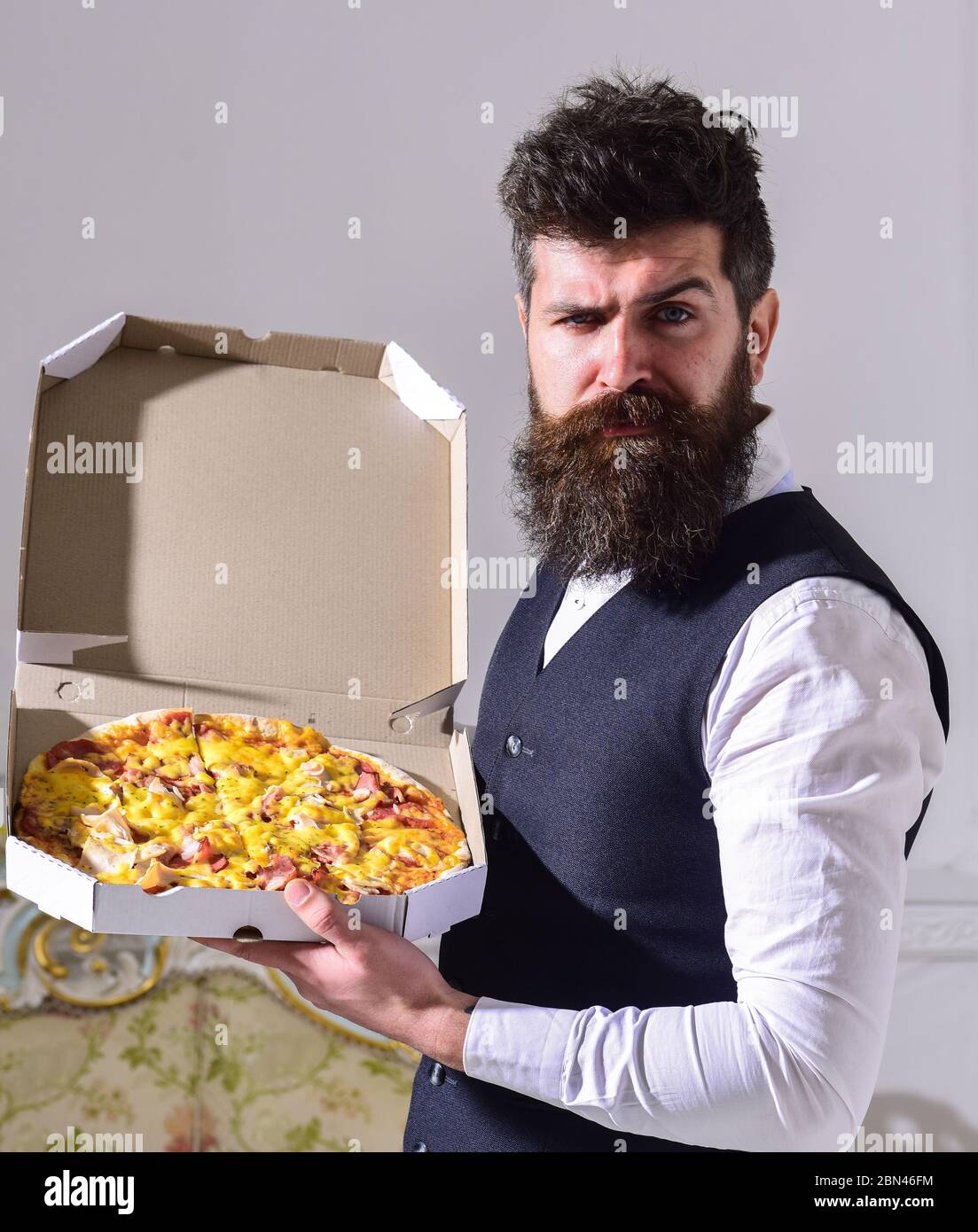 Concepto de entrega de pizza. Macho en ropa clásica hambrienta, en cara  estricta, ir a comer pizza, fondo interior de lujo. Hombre con barba y  bigote sostiene la caja entregada con deliciosa