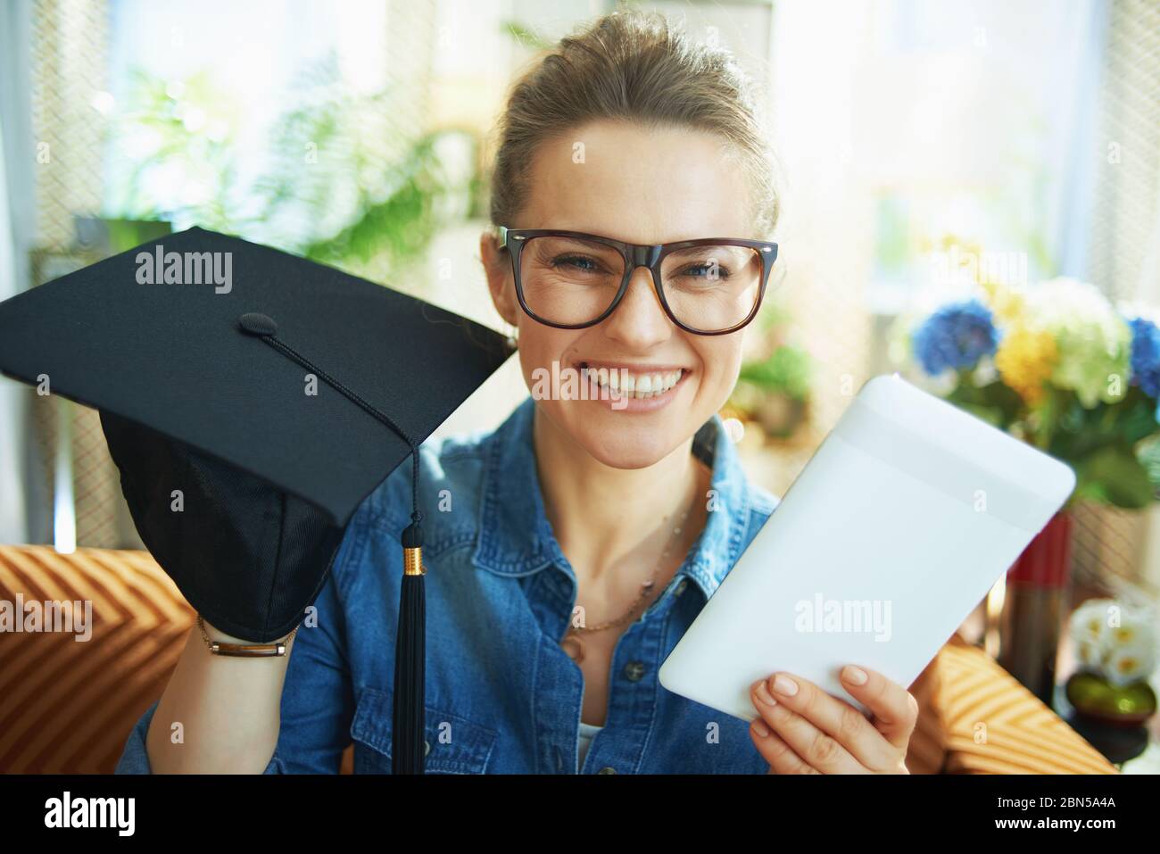 Retrato de una mujer moderna feliz en camisa de jeans con gorra de graduación y tablet PC en casa moderna en días soleados. Foto de stock