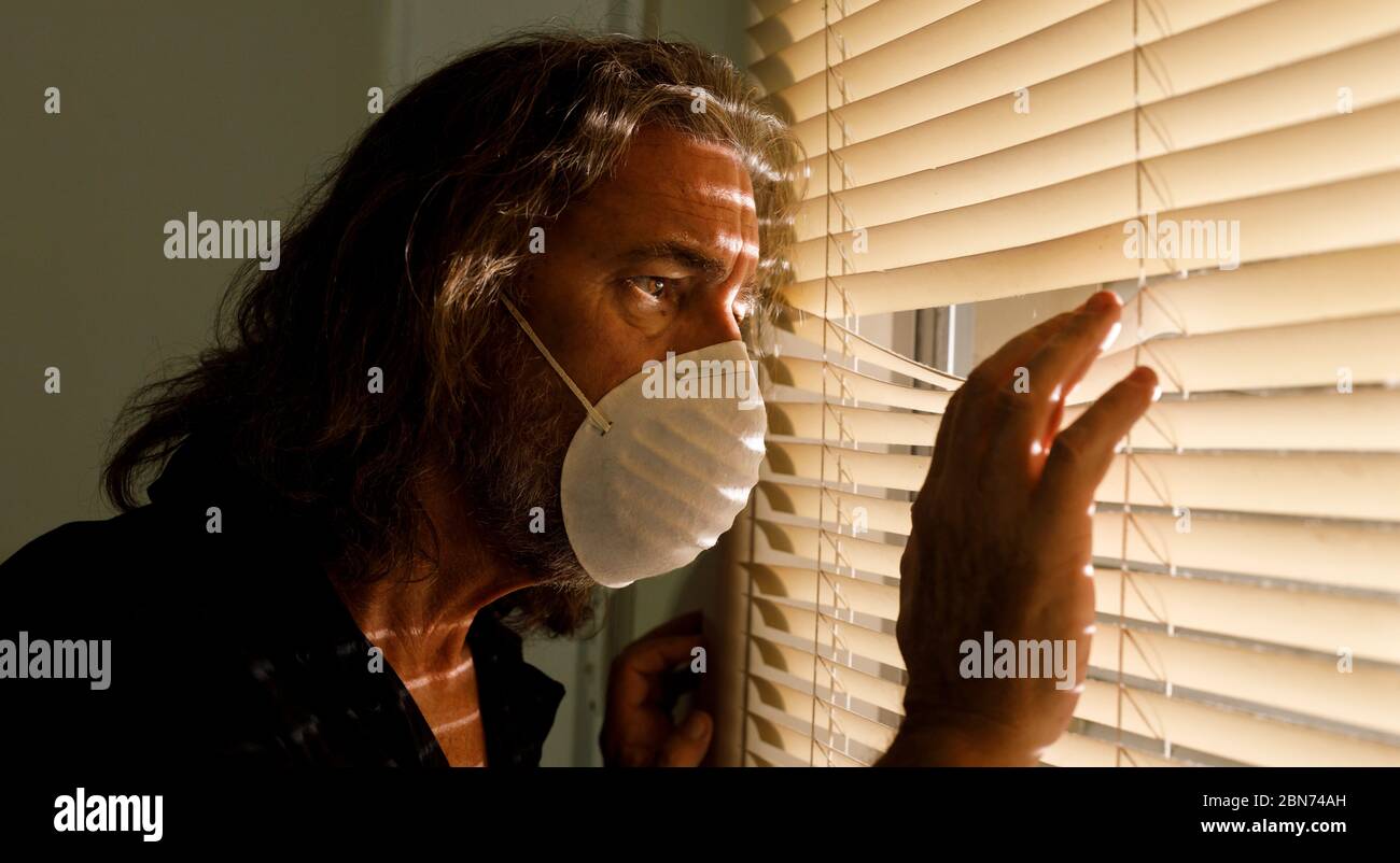 coronavirus, hombre usando máscara médica en el aislamiento de uno mismo mirando fuera de las persianas de la ventana de la casa. El hombre preocupado toma las medidas de seguridad de cuarentena de Covid 19 Foto de stock