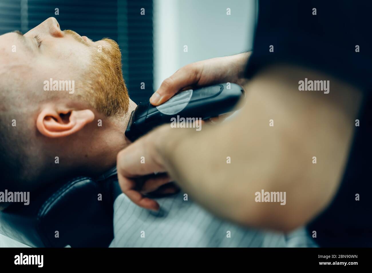 Barbería de corte barbudo hombre con máquina de afeitar en la peluquería. Proceso de peluquería. Primer plano de un estilista cortando la barba de un varón barbudo. Foto de stock