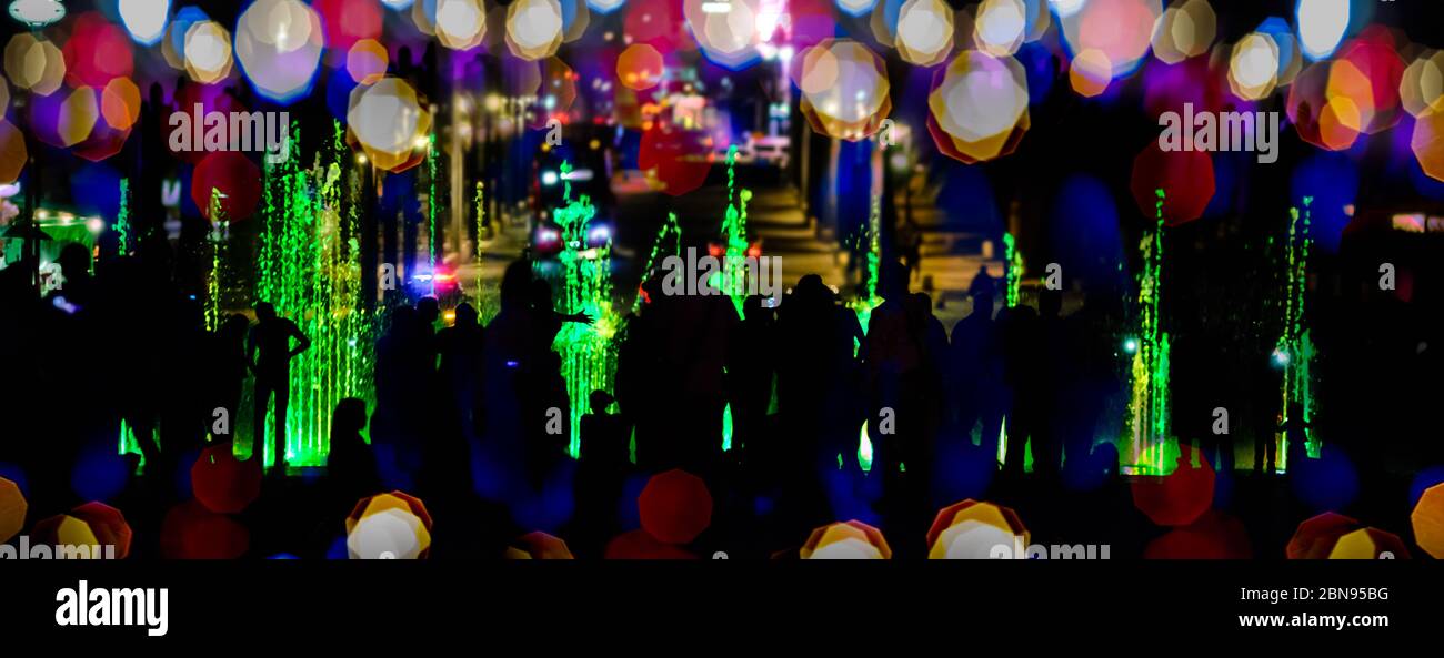 Siluetas de personas caminando y jugando en una colorida fuente iluminada por la noche Foto de stock
