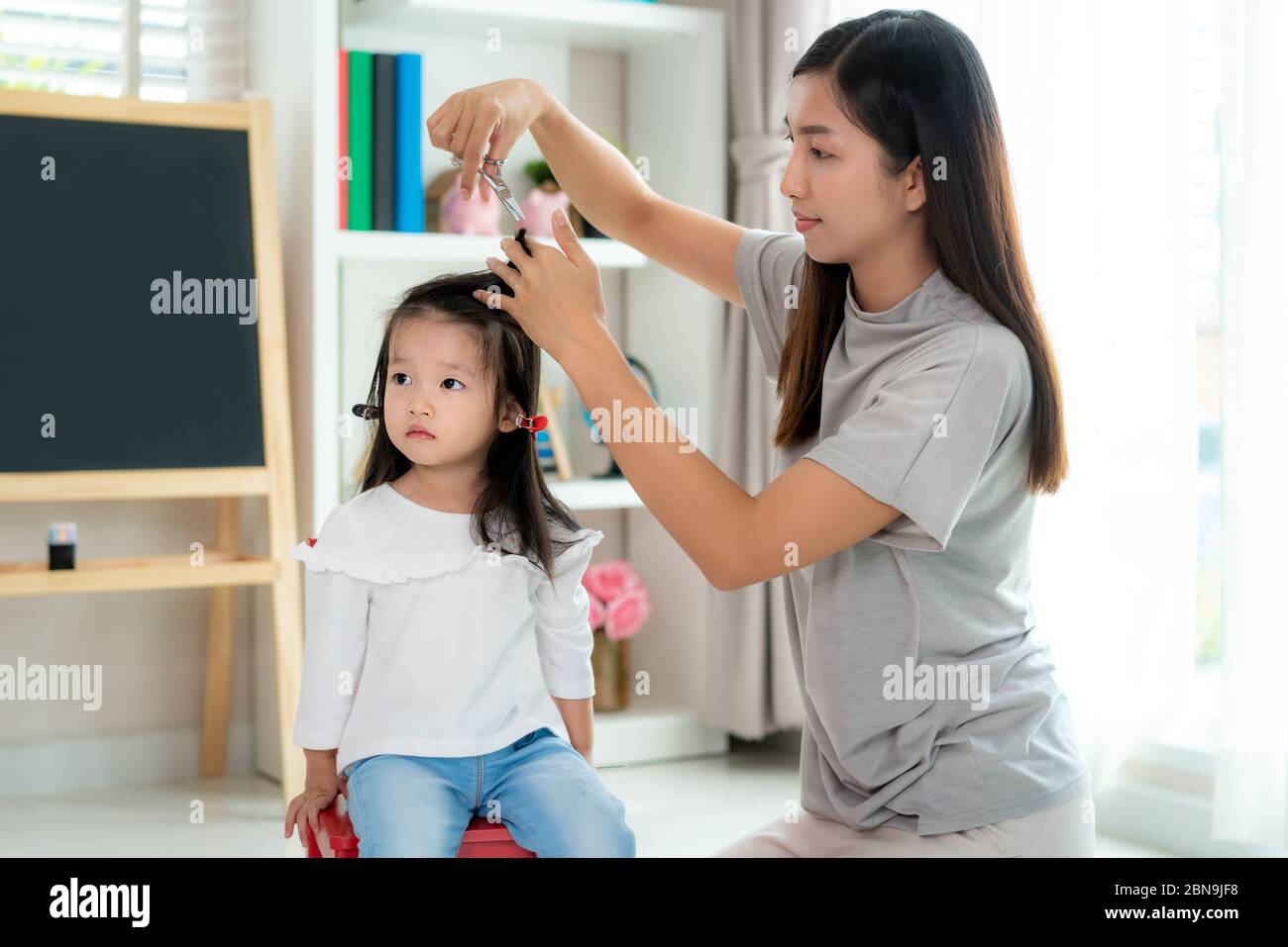 Madre Asiática cortando el pelo a su hija en la sala de estar en casa mientras se queda en casa a salvo de Covid-19 Coronavirus durante el cierre. Una cuarentena automática Foto de stock