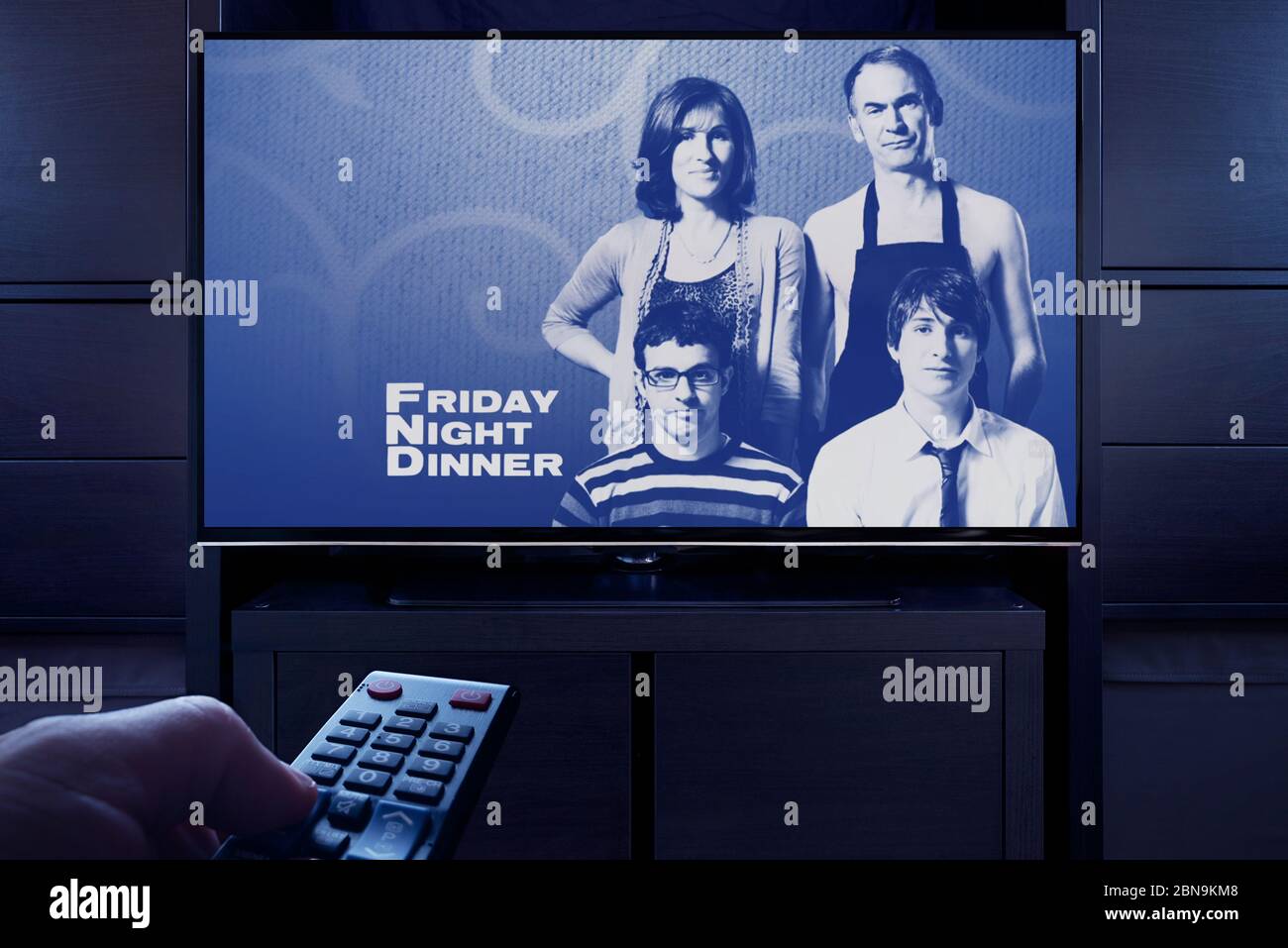 Un hombre apunta un mando a distancia de la televisión que muestra la pantalla principal del título de la cena del viernes por la noche (sólo uso editorial). Foto de stock
