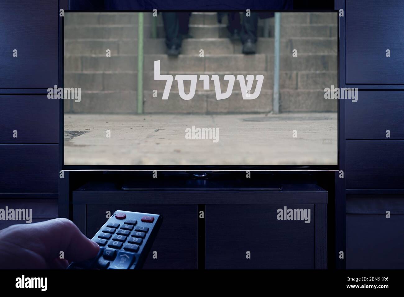 Un hombre apunta un mando a distancia de la televisión al televisor que muestra la pantalla principal del título de Shtisel (solo para uso editorial). Foto de stock