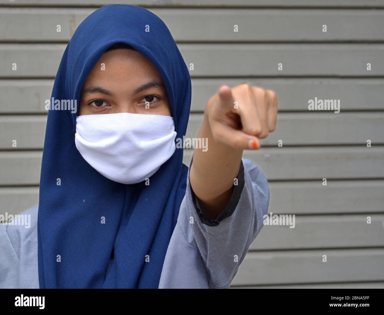 Chica musulmana malaya con hijab y puntos de velo que cubren la boca durante la pandemia de Corona con su dedo índice hacia el espectador: ¡mantenga su distancia social! Foto de stock