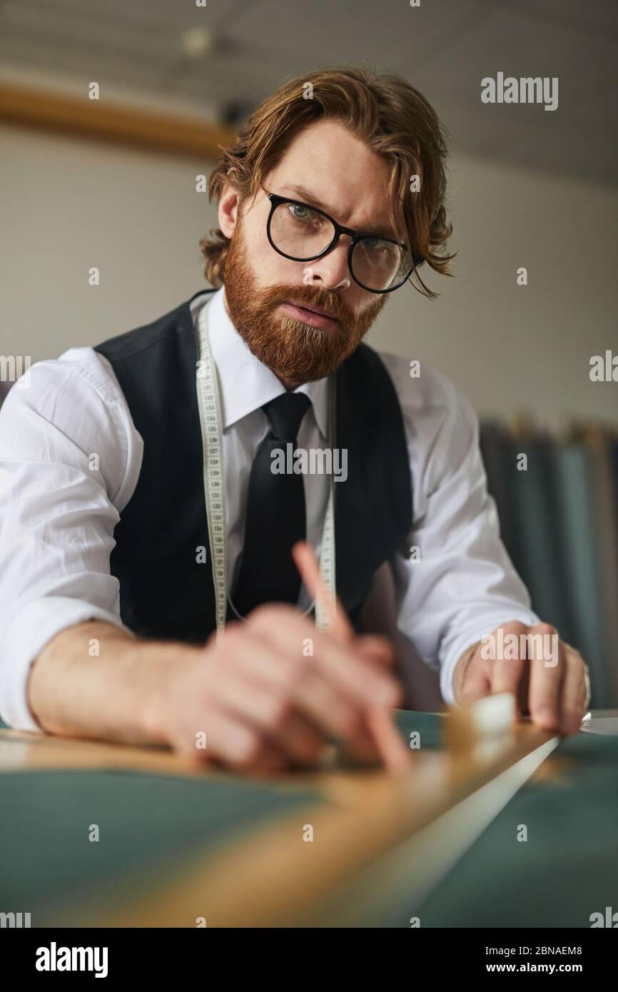 Retrato de sastre con barba en gafas mirando la cámara mientras se trabaja con tela en la mesa Foto de stock