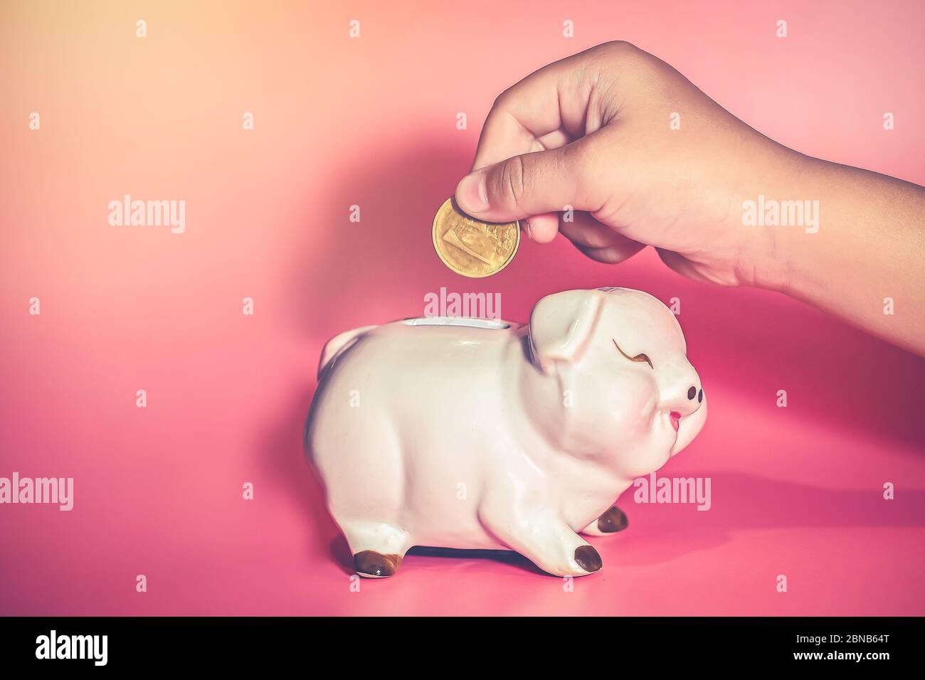 La mano de niño soltar una moneda en Piggy Bank para ahorrar con montón de monedas en la mesa en casa, ahorrar dinero para el aprendizaje futuro. Foto de stock