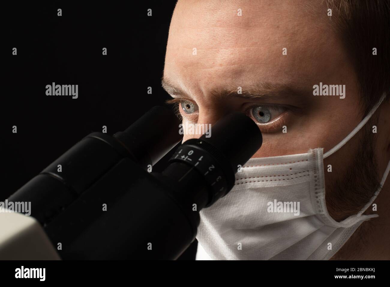 Foto de cerca de un científico masculino blanco usando un microscopio binocular y usando una máscara facial. Foto de stock