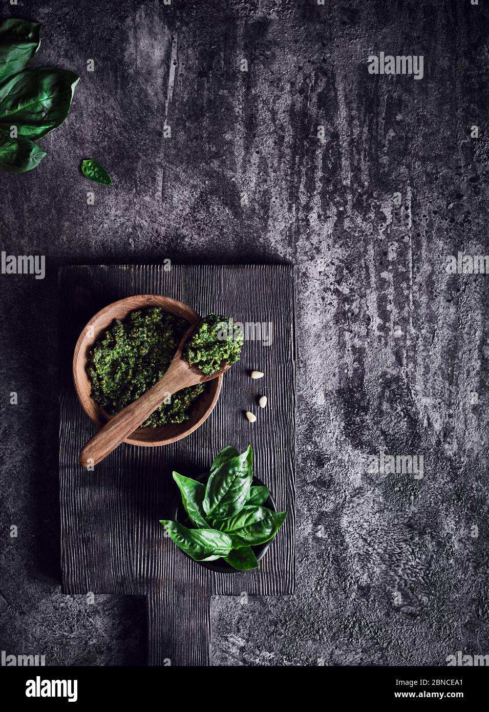 Salsa pesto de albahaca verde, parmesano, ajo, piñones en un tazón rústico de madera con cuchara sobre fondo texturizado oscuro Foto de stock