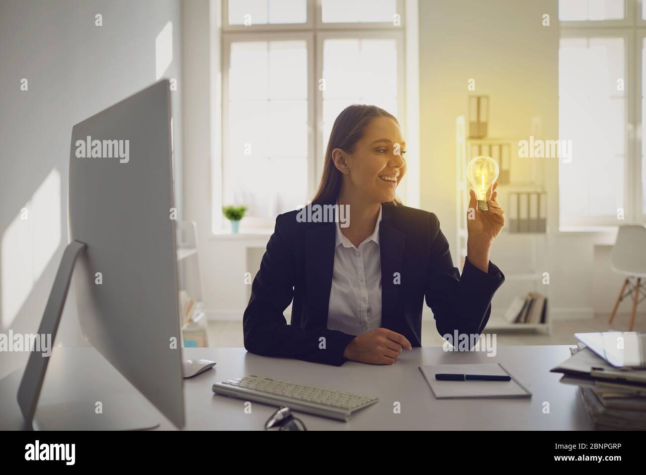 Feliz mujer de negocios inteligente sosteniendo la bombilla de la luz ardiente que tiene una idea brillante mientras que trabaja en la oficina Foto de stock