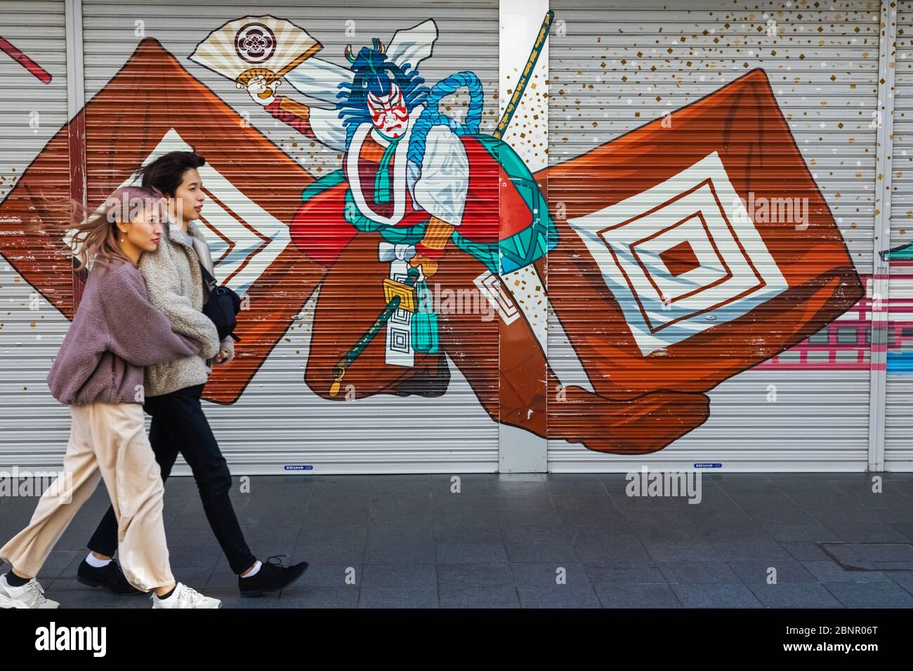 Japón, Honshu, Tokio, Asakusa, Pareja Joven Caminando Frente A La Colorida Tienda De Pintura Obturador Que Representa La Escena Kabuki Foto de stock