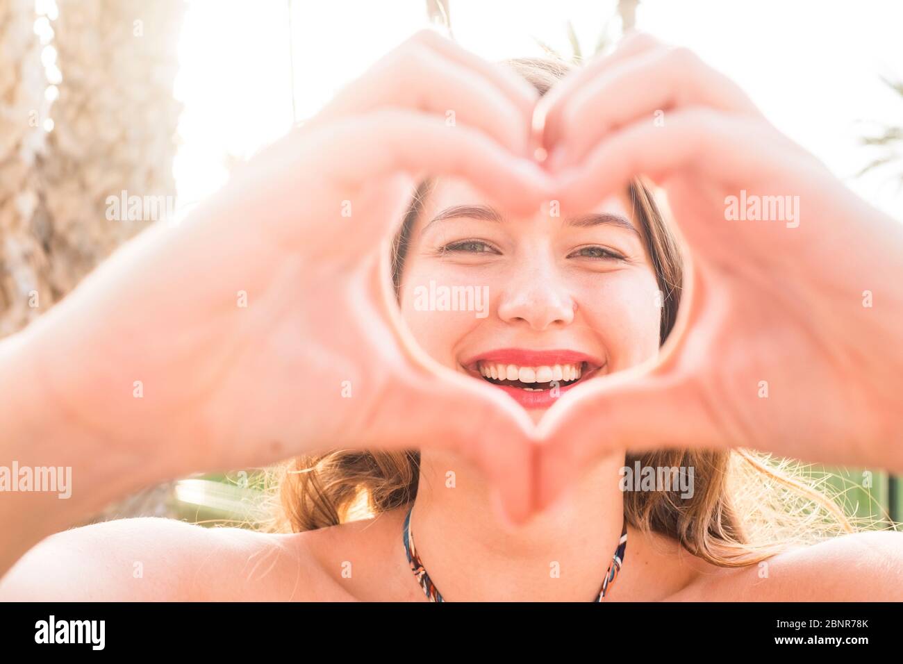 Retrato de cerca de hermosa. Mujer joven caucásica haciendo amor signo de corazón con las manos en la cámara - sol brillante fondo y felicidad alegre gente concepto Foto de stock