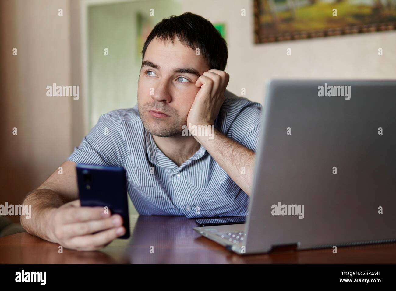 Triste lindo joven sosteniendo un smartphone en su mano un hombre trabajando en un portátil de forma remota. Fatiga por cuarentena, auto-aislamiento. Foto de stock