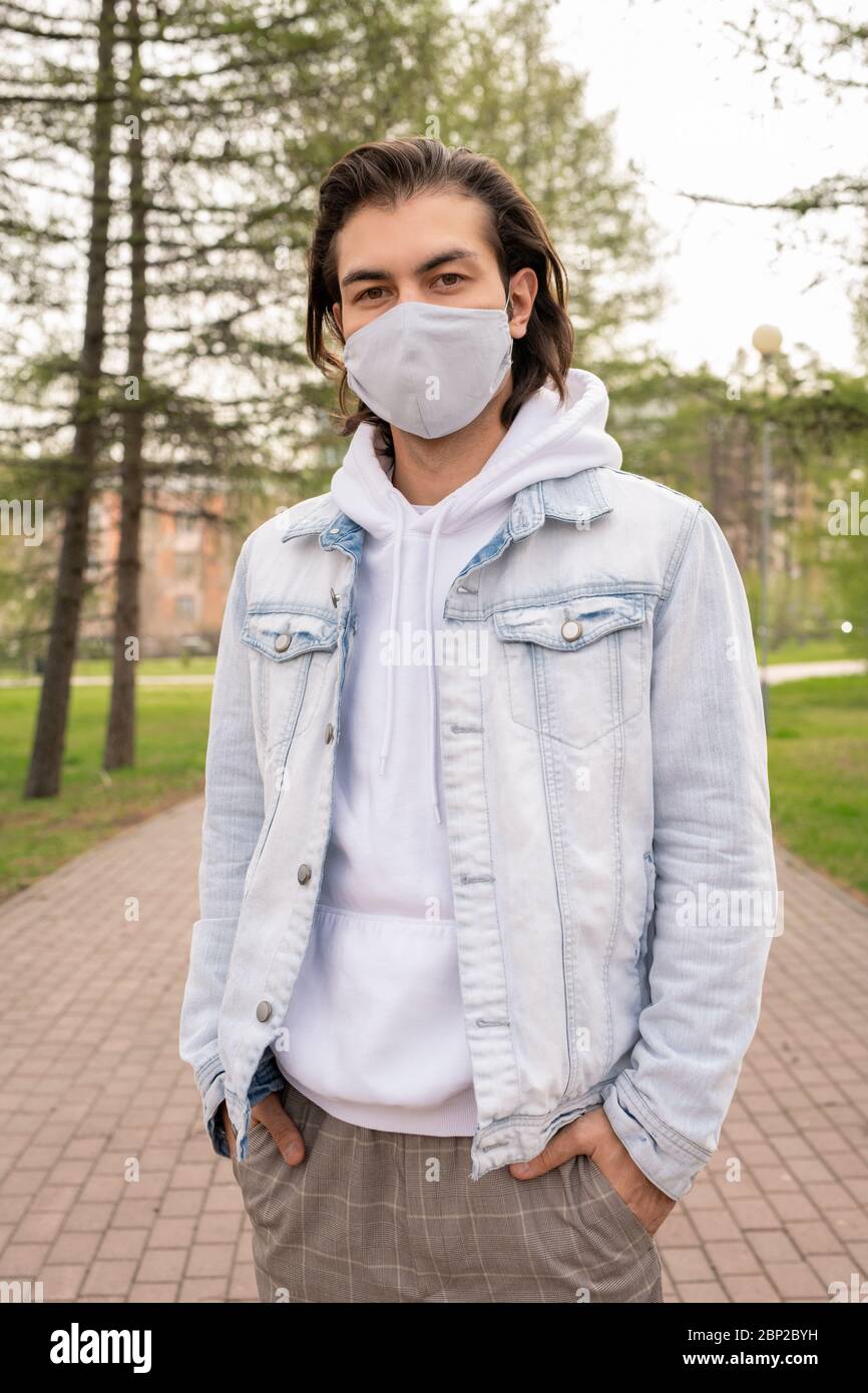 Joven en máscara protectora, sudadera con capucha blanca y chaqueta ligera  de tela vaquera que mantiene las manos en bolsillos de pantalones mientras  está de pie en la carretera del parque público