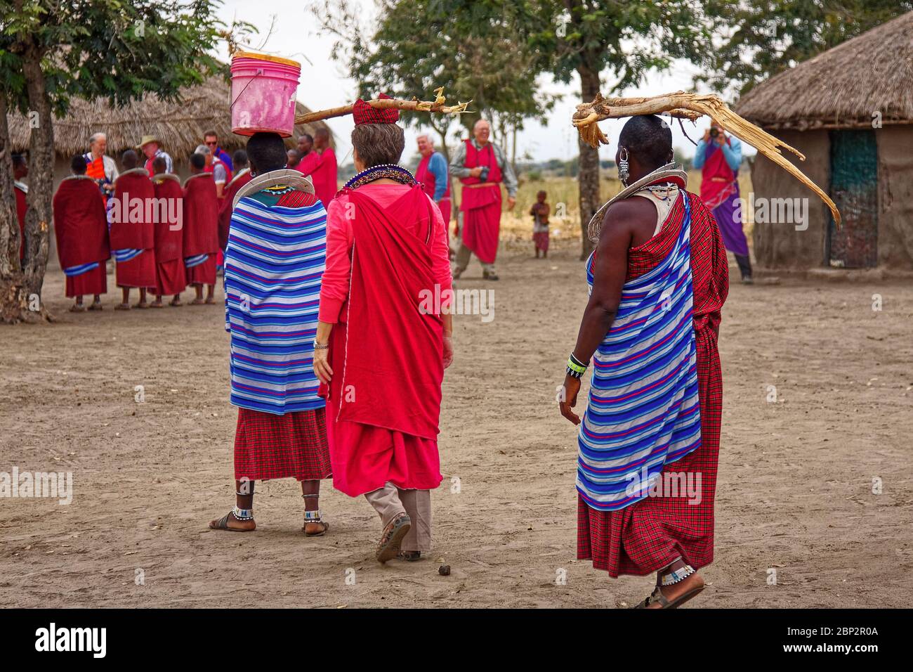 Aldea Maasai; 3 mujeres llevando objetos en la cabeza, 2 Massai, 1 visitante, nativos; vestido tradicional, aprendizaje, educación, compartir cultura, volver vi Foto de stock