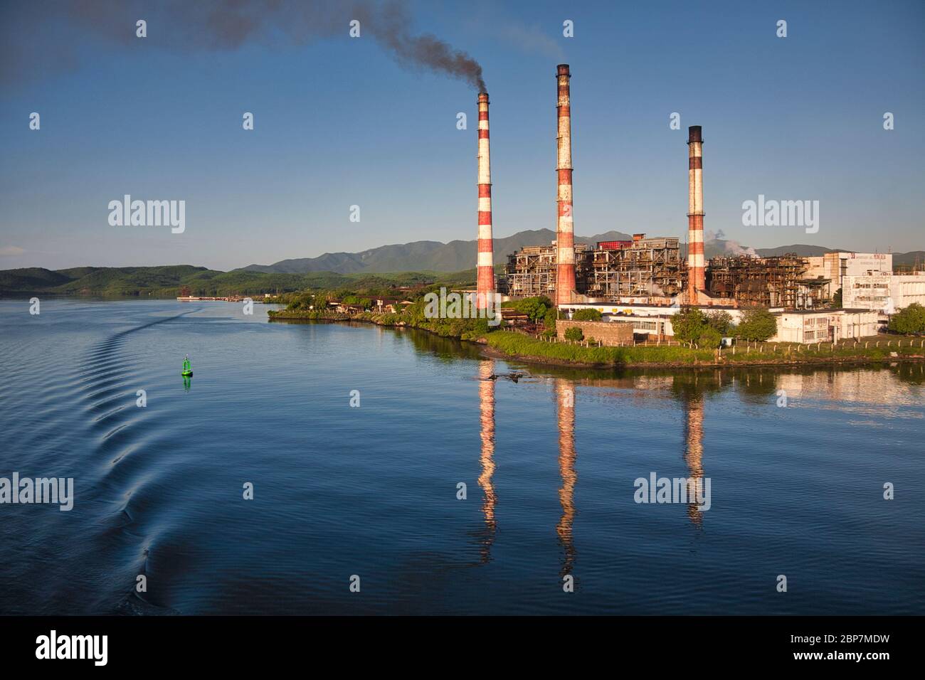 Santiago de Cuba en la costa sur de Cuba. Una central eléctrica con tres chimeneas reflejadas en el agua de la entrada en la que se encuentra Foto de stock
