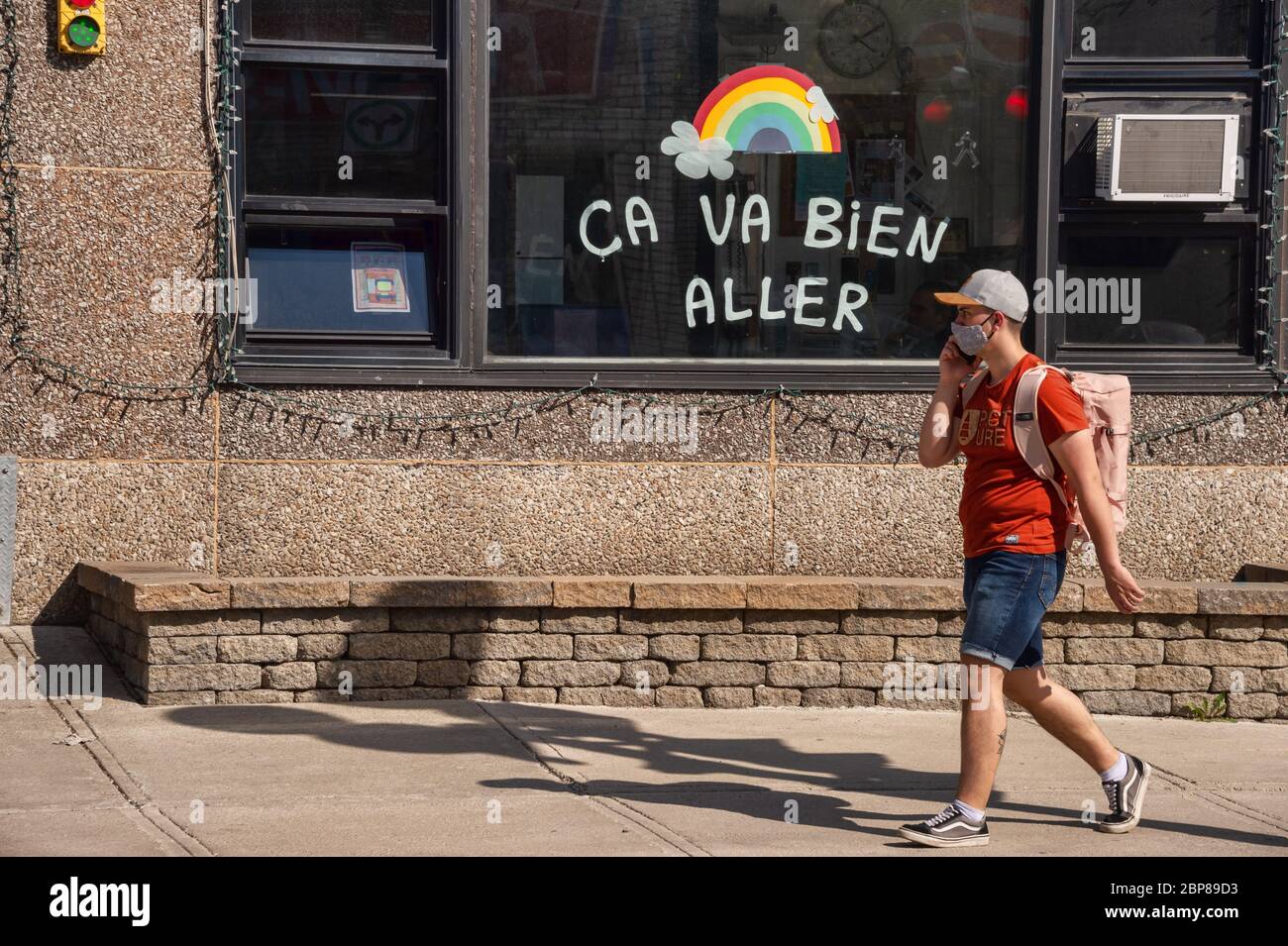 Montreal, CA - 17 de mayo de 2020: Joven con máscara para la protección contra COVID-19 montando una bicicleta frente al arco iris dibujando en la calle Masson. Foto de stock