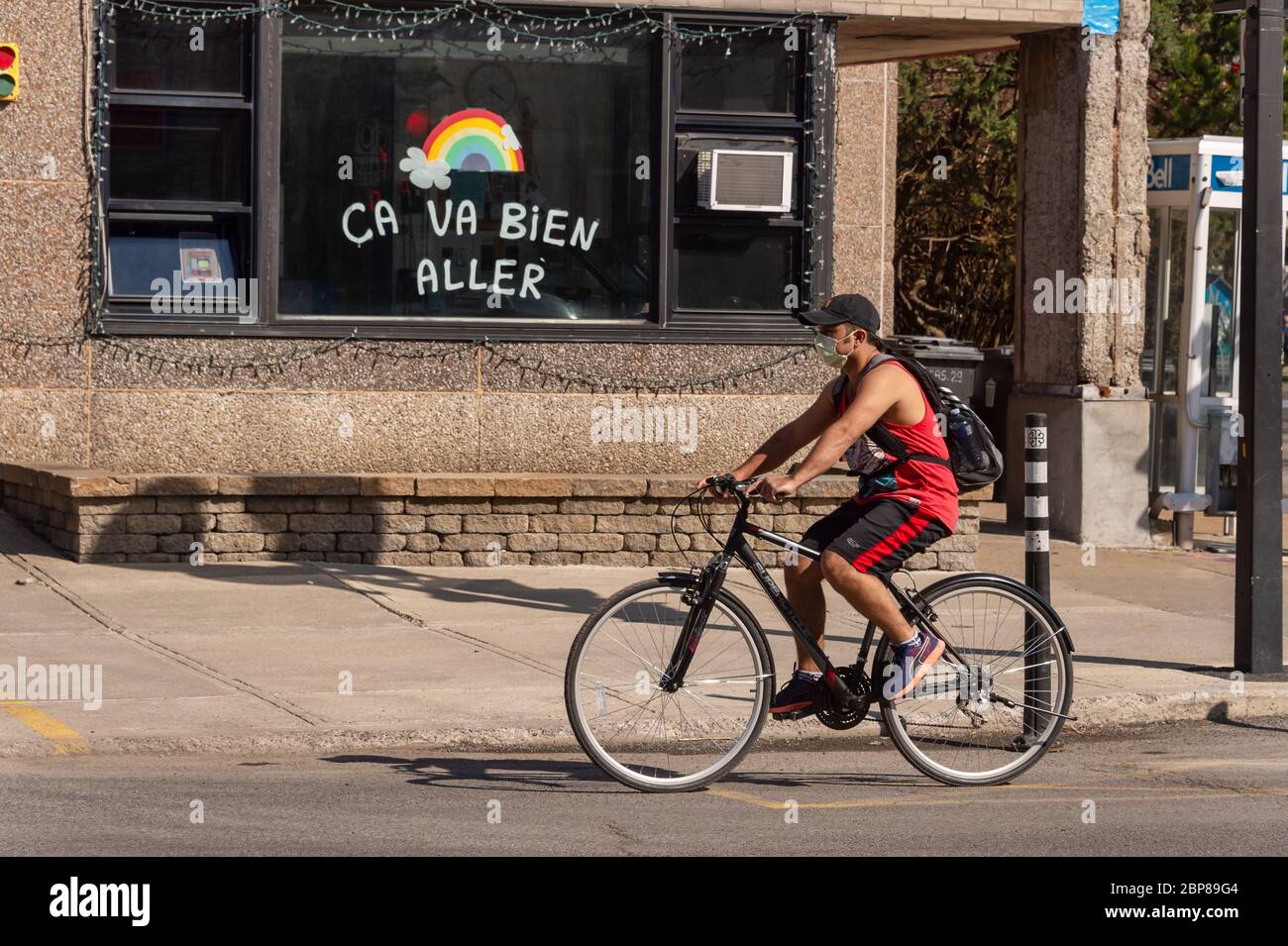 Montreal, CA - 17 de mayo de 2020: Joven con máscara para la protección contra COVID-19 montando una bicicleta frente al arco iris dibujando en la calle Masson. Foto de stock