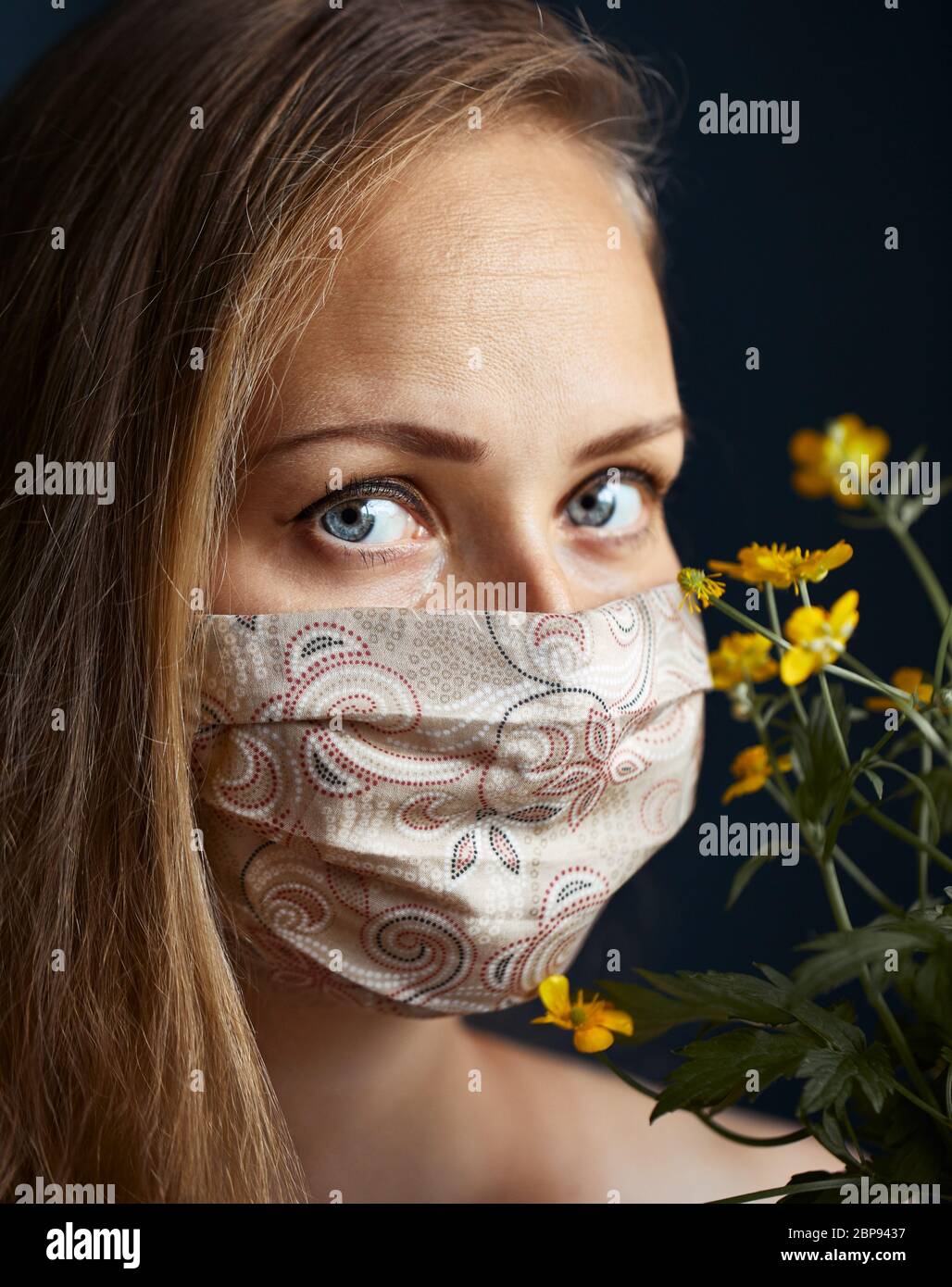 Mujer hermosa con una máscara floral hecha a mano cara, mirando la cámara, cerca de fondo oscuro. Epidemia de gripe, alergia al polvo, protección contra vi Foto de stock